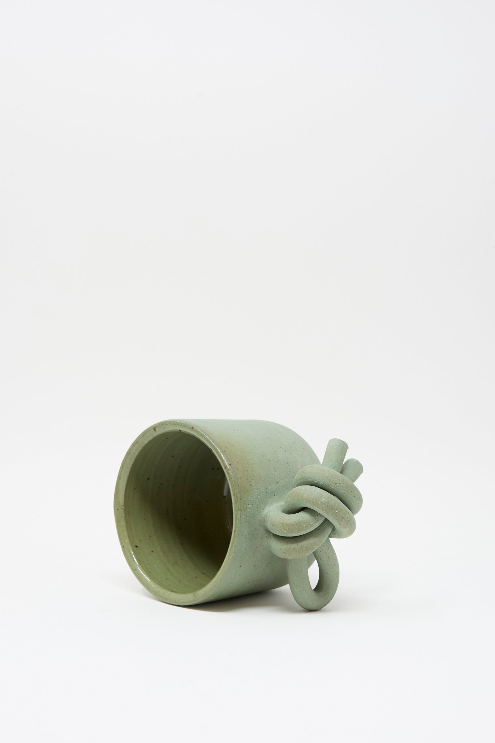 The Hand Built Knot Mug in Sage I by Lost Quarry, a green handmade ceramic mug with a distinctive oversized twisted and knotted handle, is shown lying on its side against a plain white background.
