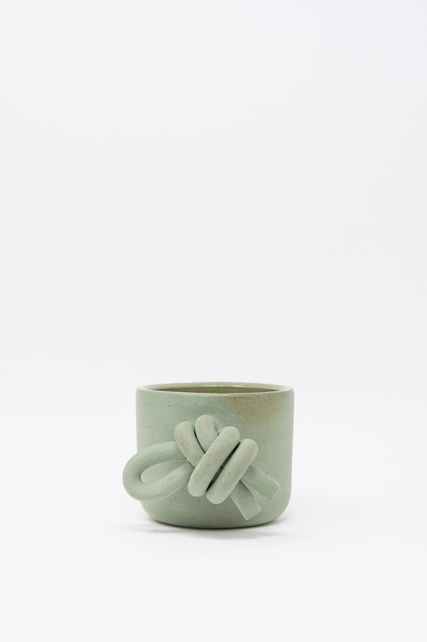 The Lost Quarry Hand Built Knot Mug in Sage I is a small, matte light green ceramic mug with a decorative knot-shaped oversized handle, shown on a plain white background.