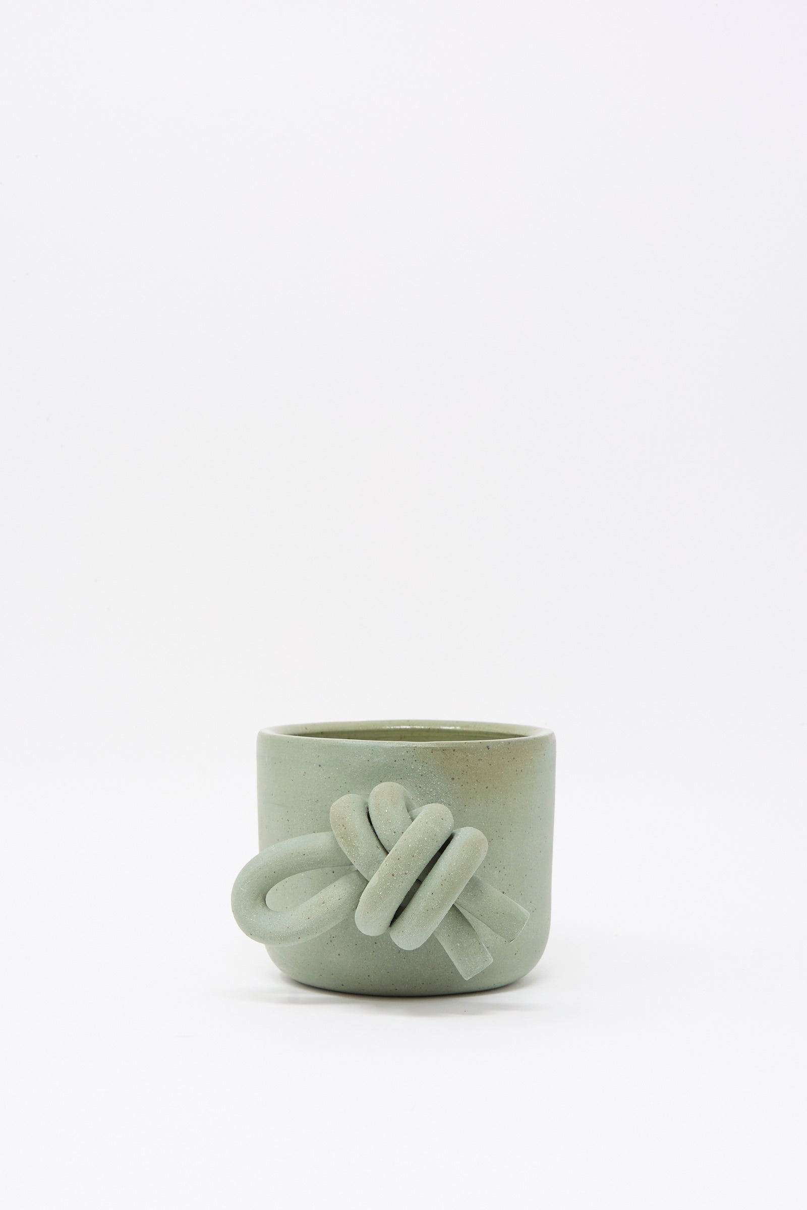 The Lost Quarry Hand Built Knot Mug in Sage I is a small, matte light green ceramic mug with a decorative knot-shaped oversized handle, shown on a plain white background.