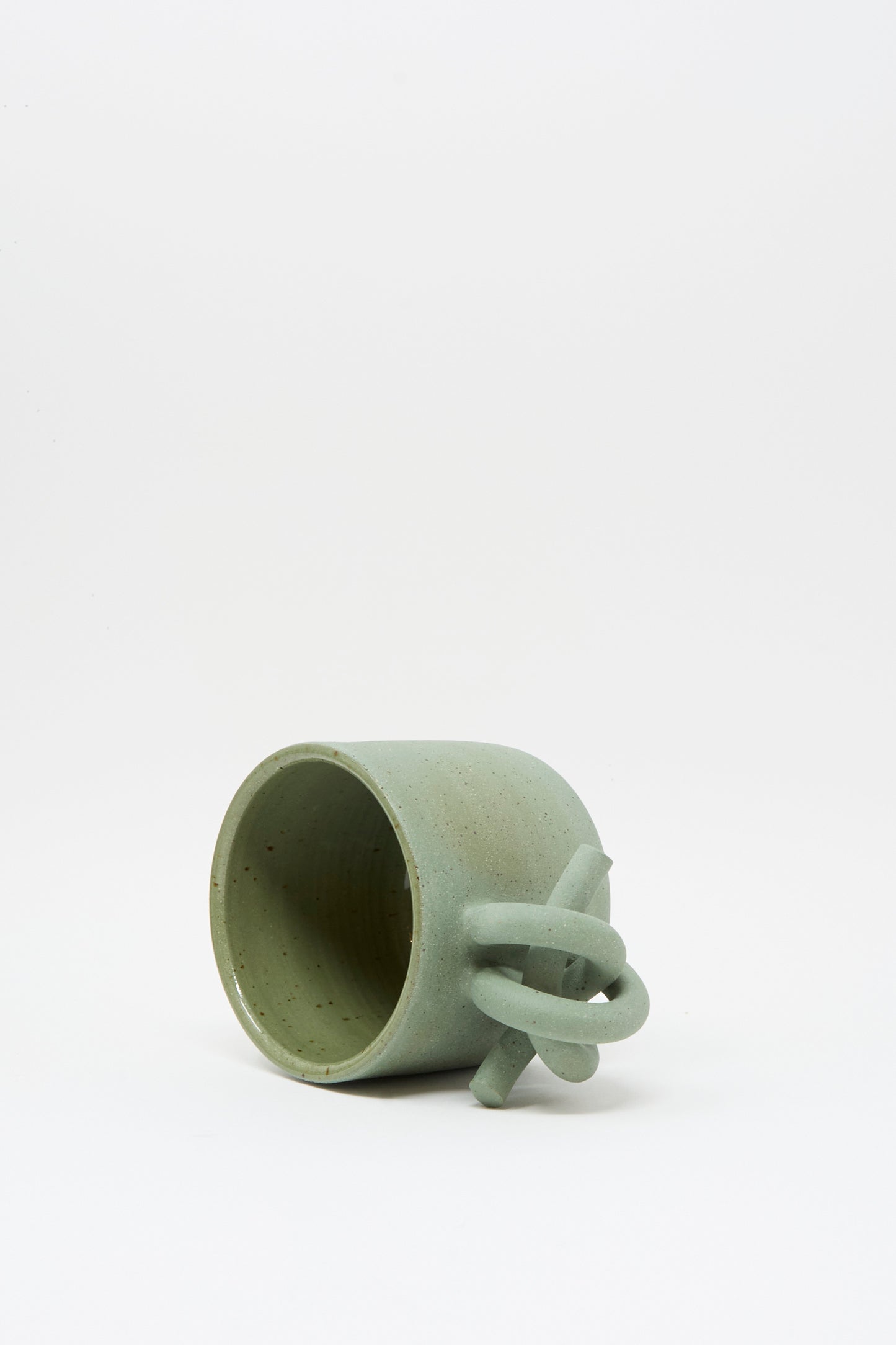 The Lost Quarry Hand Built Knot Mug in Sage II is a handmade green ceramic mug with a unique looped handle and soft sage glaze, shown tipped on its side against a plain white background.