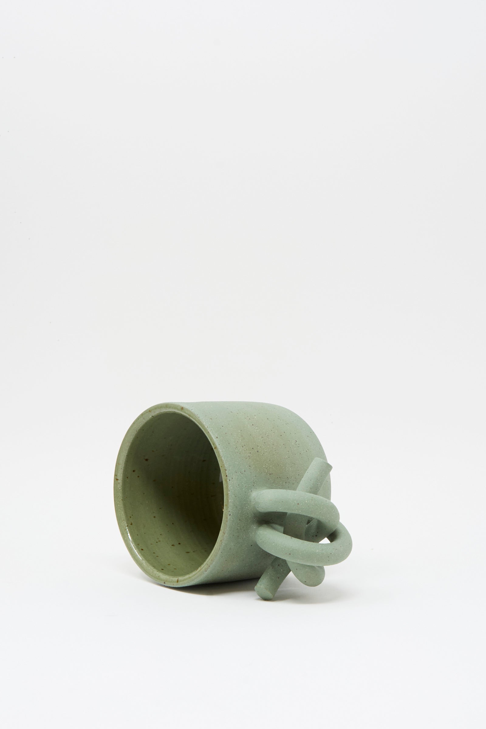 The Lost Quarry Hand Built Knot Mug in Sage II is a handmade green ceramic mug with a unique looped handle and soft sage glaze, shown tipped on its side against a plain white background.