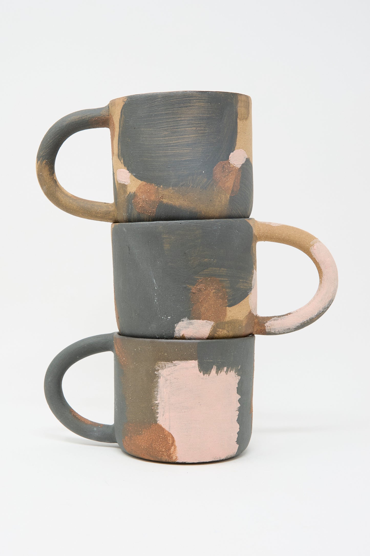 Three ceramic mugs with abstract designs in earthy brown, gray, and pink tones, Lost Quarry's Hand Built Mugs in Brown Multi, are stacked against a white backdrop.