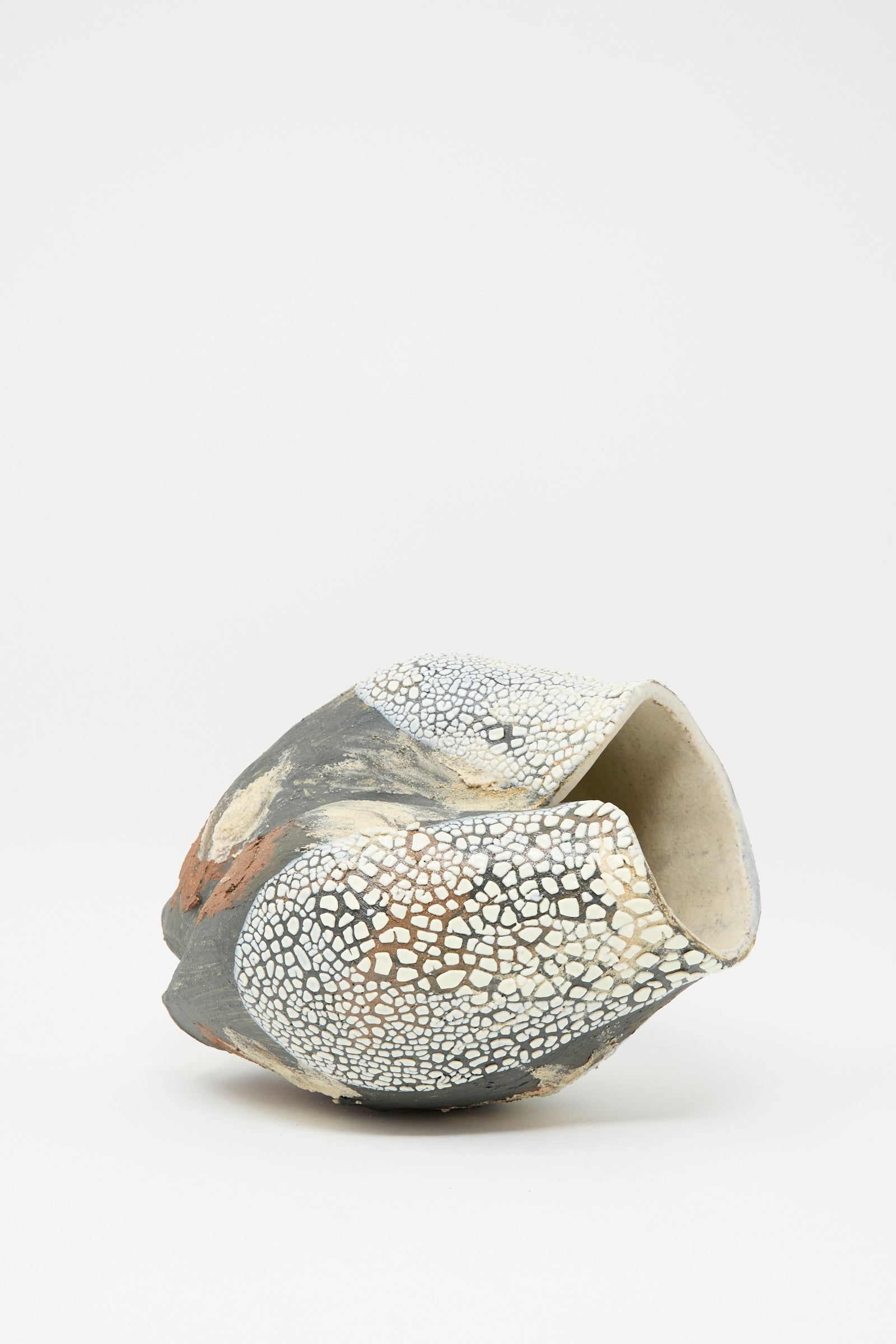 The Lost Quarry Hand Built Textured Vessel in Black Multi features a sculptural design with a rough, cracked texture, blending dark gray, white, and terracotta lays on its side on a plain white backdrop.