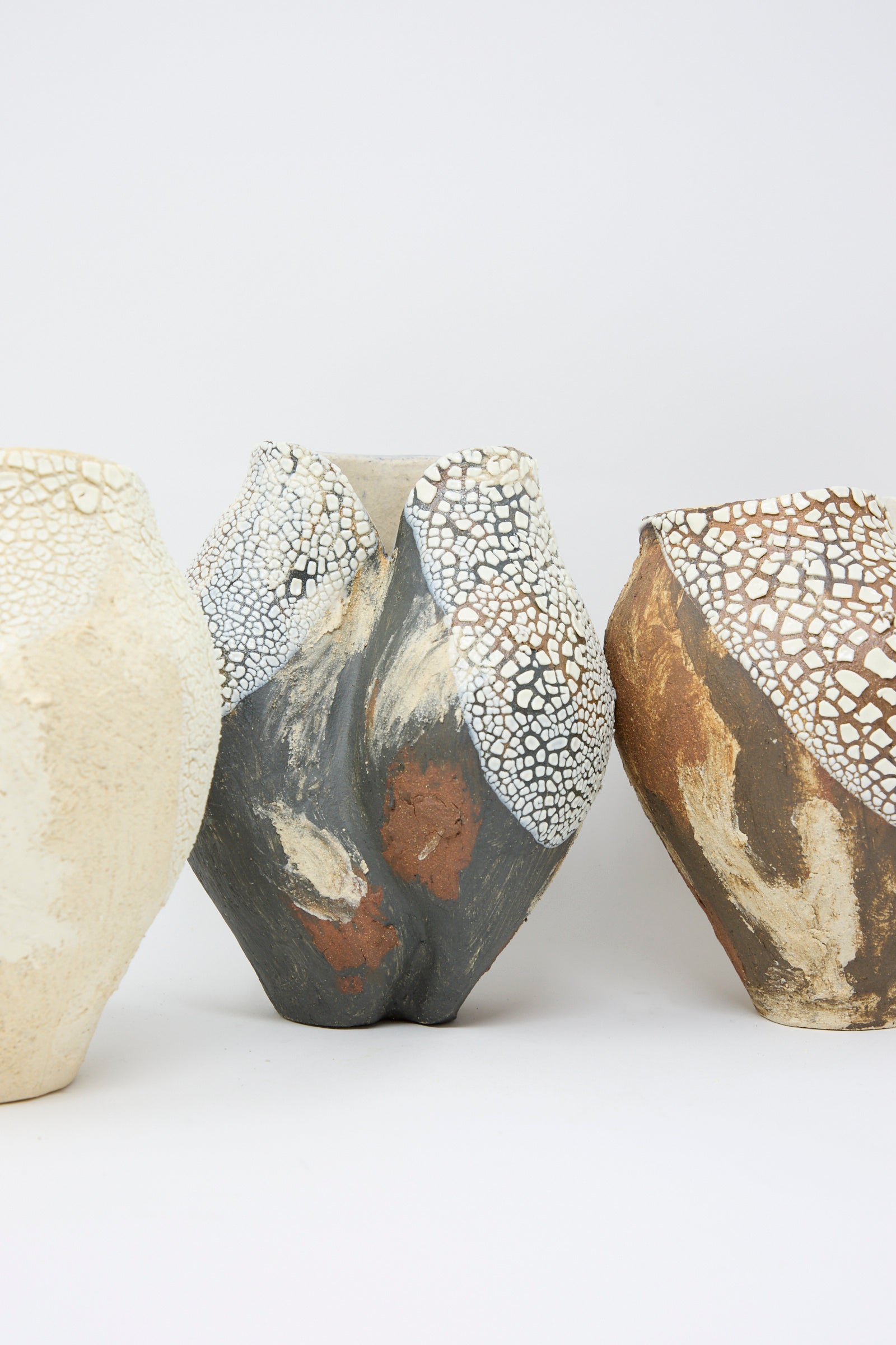 Lost Quarry's Hand Built Textured Vessel in Black Multi, along with two others, showcases sculptural clay forms and textured, cracked surfaces in cream, gray, and brown tones against a plain background.