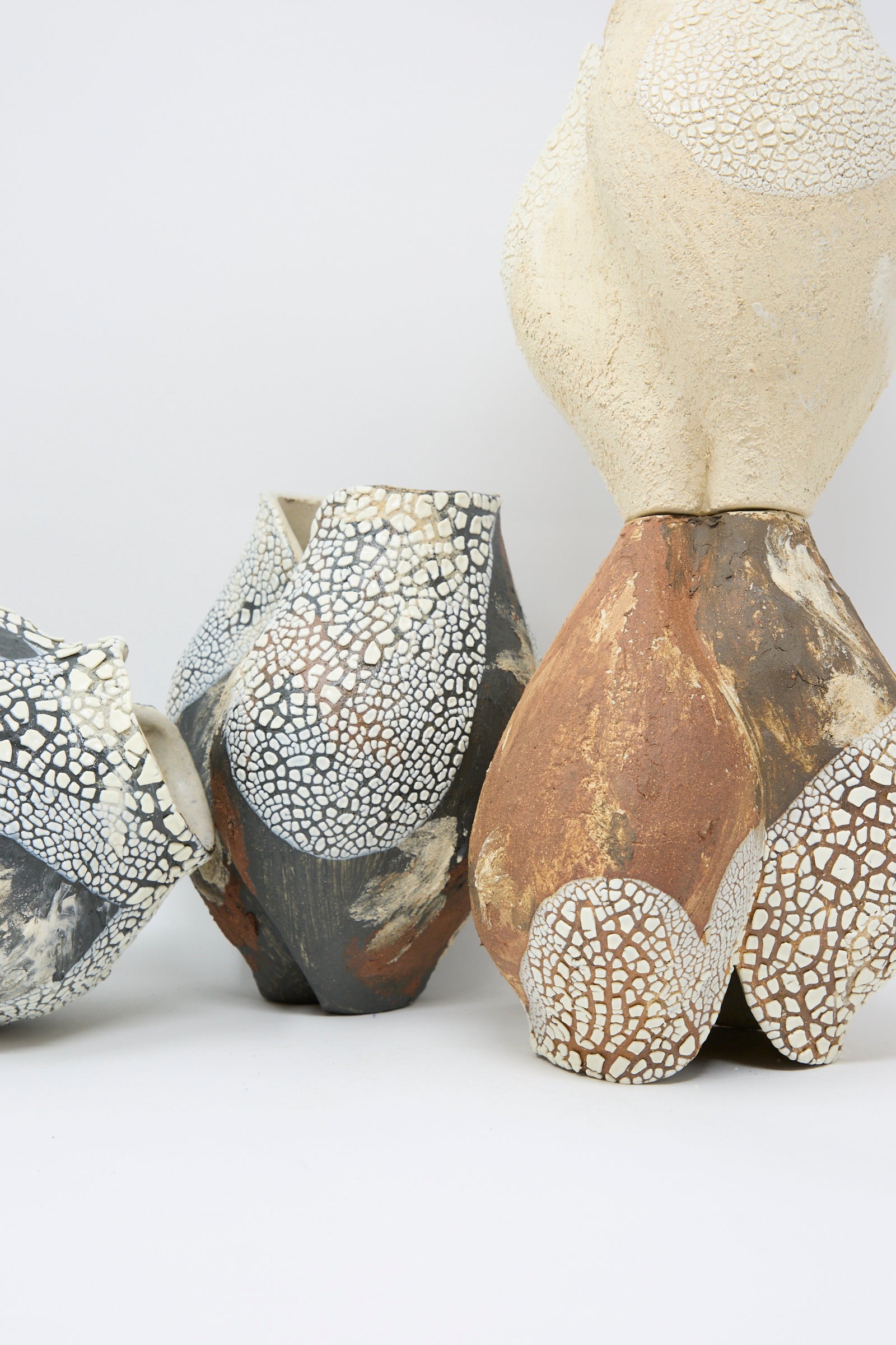 The Lost Quarry Hand Built Textured Vessel in Terracotta Mix is shown along with three other abstract ceramic sculptures with a textured glaze and cracked surfaces in shades of gray, white, and tan, displayed against a plain white background.