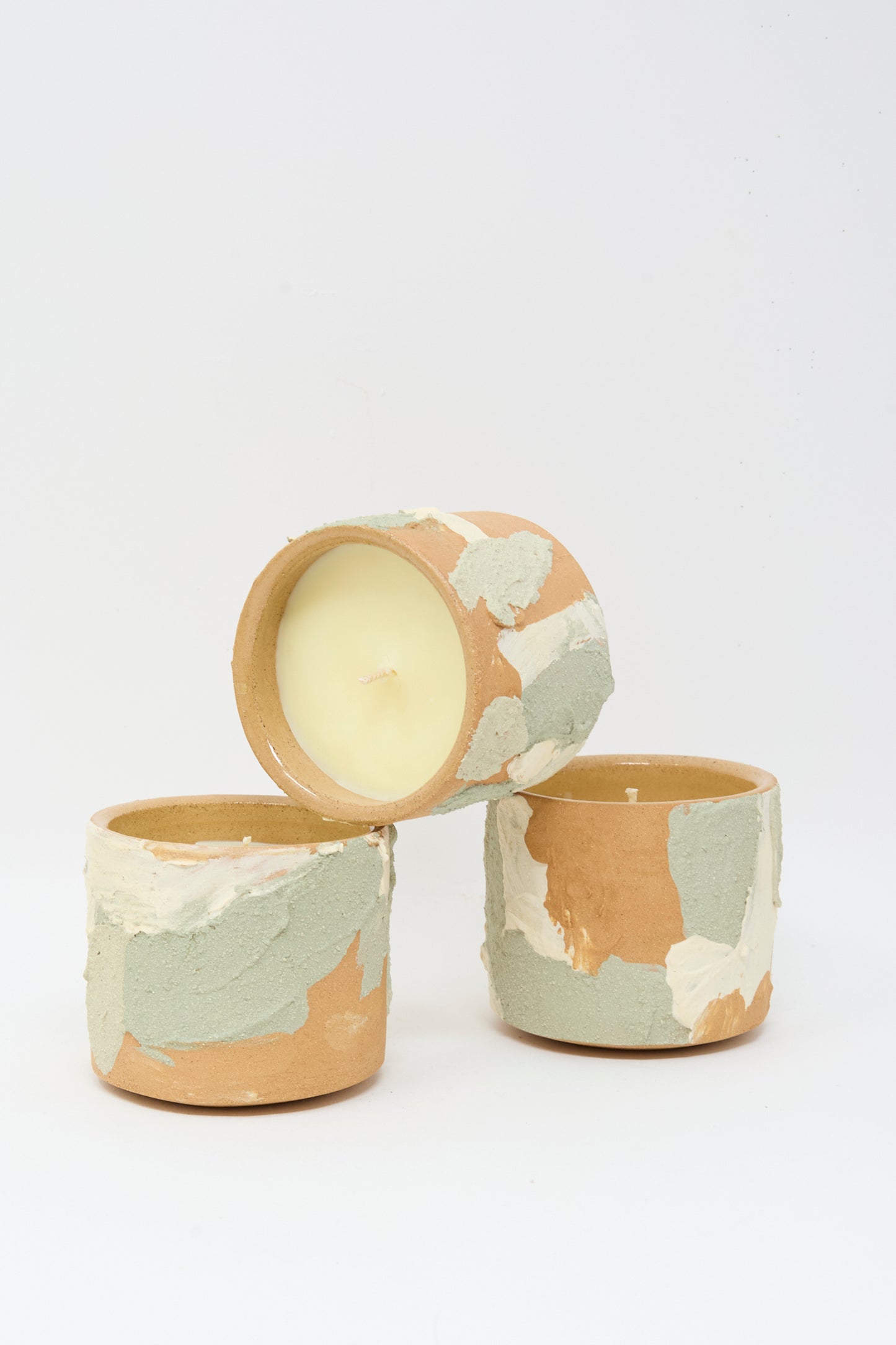 Three Neroli candles by Lost Quarry, in textured, earth-toned Dusty Mint Adobe Multi ceramic vessels, are arranged triangularly—with one candle resting horizontally atop two upright holders.