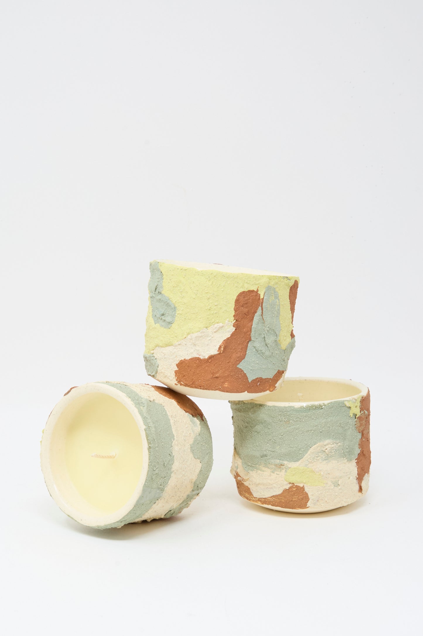 Three Neroli Candles in Lime Terracotta Multi by Lost Quarry, each in hand-built ceramic vessels with a green, beige, and brown camouflage pattern, are arranged on a white background. One candle lies on its side to reveal the wick.