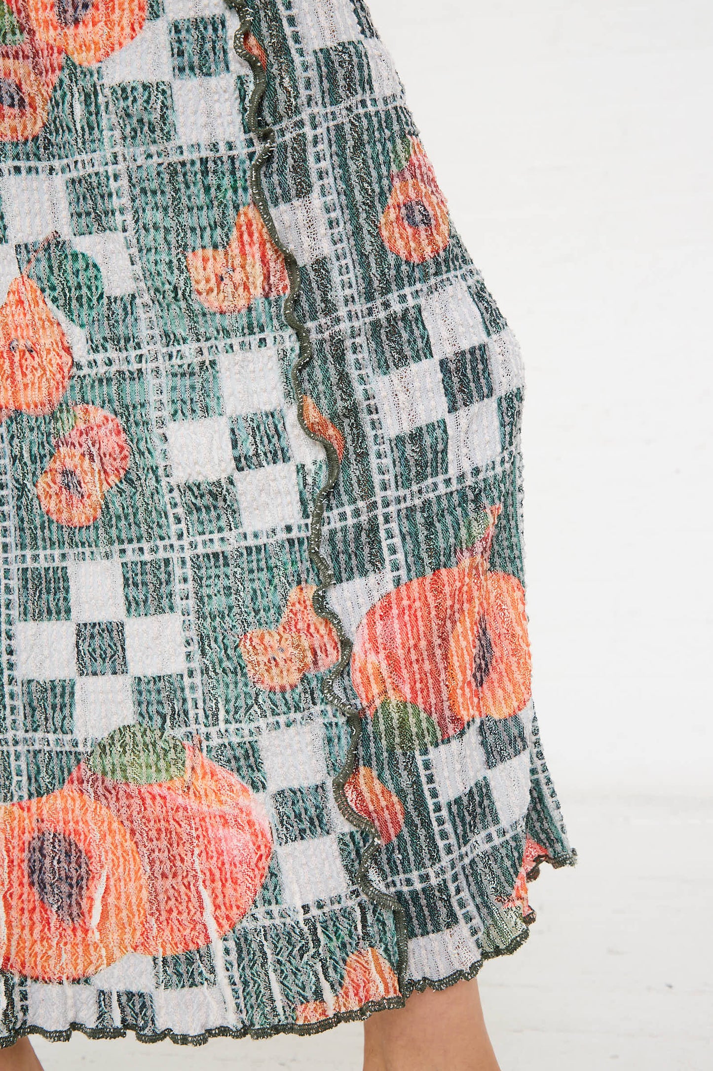 Close-up of the El Mantel Stretch Weave Luna Dress in Mandarin Tree by Luna Del Pinal, showcasing a patterned fabric with a checkered design featuring green, white, and orange floral elements. The fabric is detailed with textured, ruffled edges and evokes the charm of a stretch maxi dress.