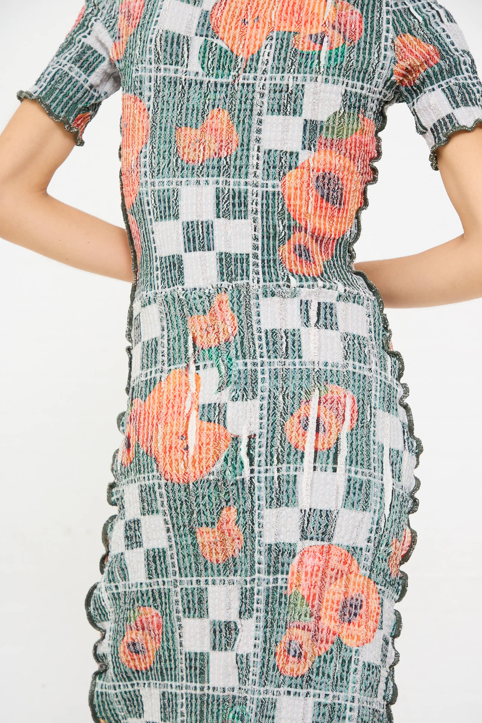 A close-up view of a person wearing the Luna Del Pinal "El Mantel Stretch Weave Luna Dress in Mandarin Tree," featuring a checkered pattern, orange floral designs, and scalloped edges on the sleeves and seams.