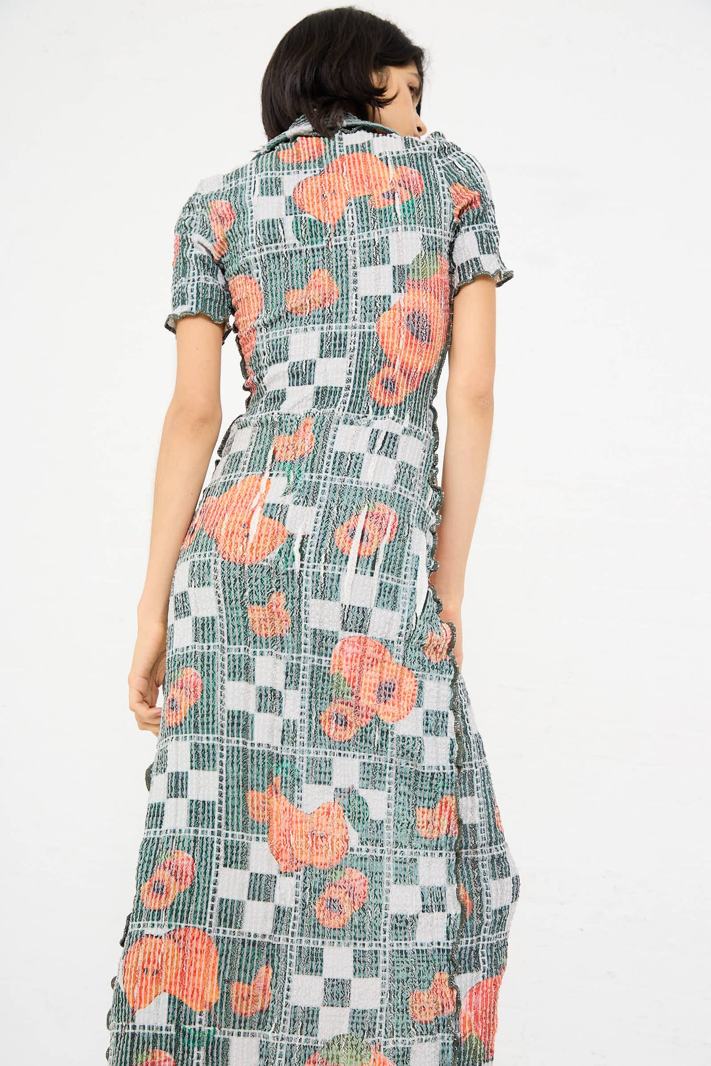 A person with short dark hair is standing with their back to the camera, wearing the El Mantel Stretch Weave Luna Dress in Mandarin Tree by Luna Del Pinal, which features a checked pattern and large red floral designs.