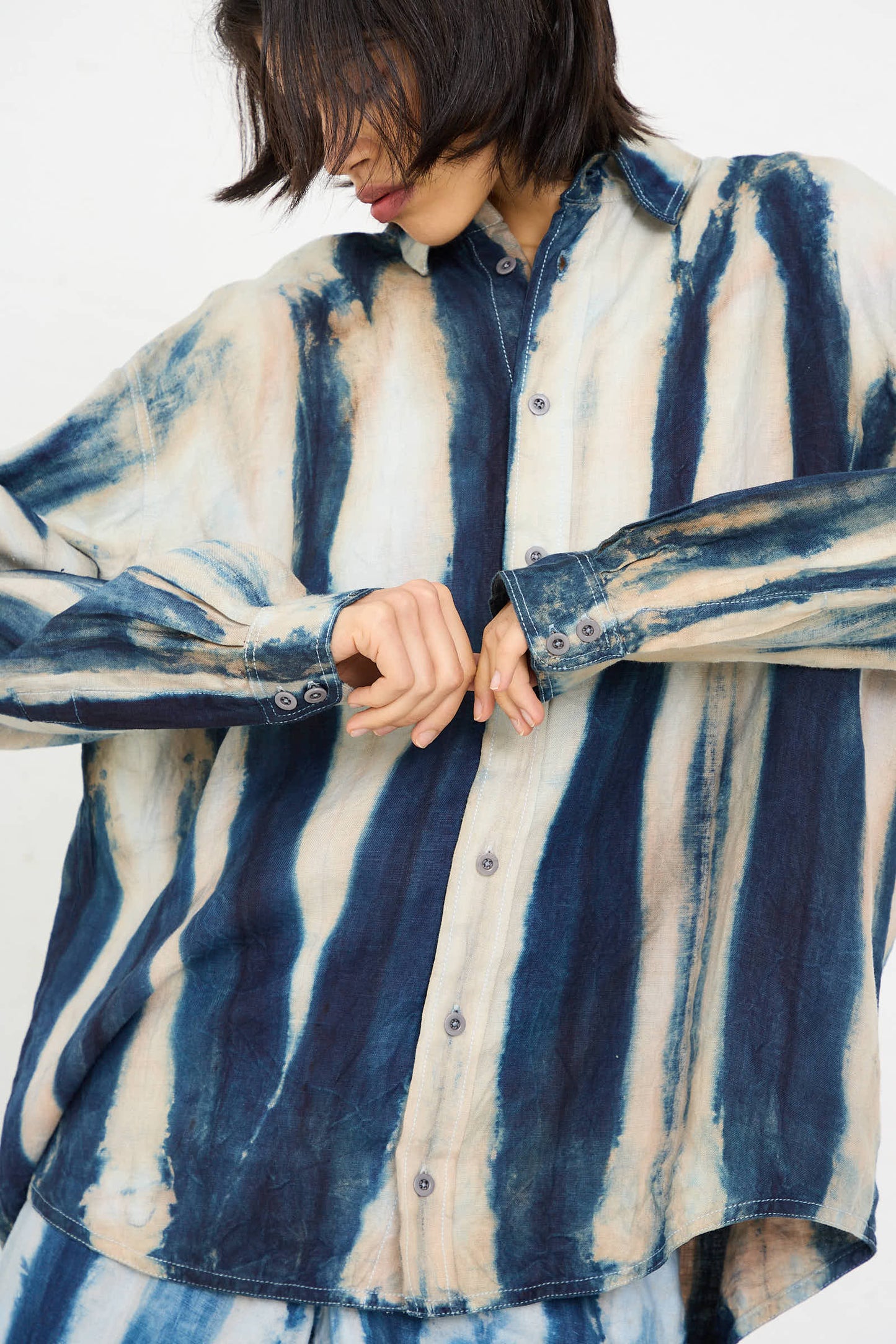 Person wearing a Shibori Hemp Overshirt in Faded Indigo by Luna Del Pinal, adjusting the cuffs of the oversized, long-sleeved shirt adorned with vertical faded indigo stripes. Their face is partially obscured by their hair. The fabric of the shirt is made from sustainably grown hemp.