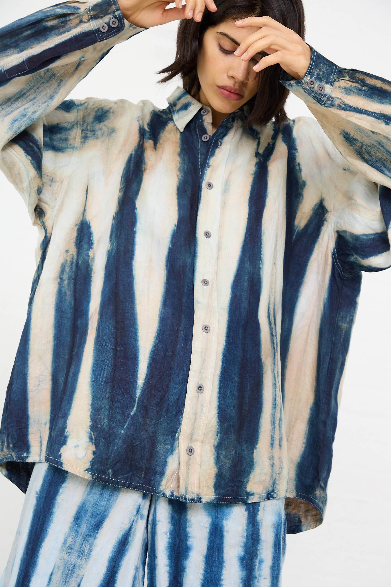 A person wearing Luna Del Pinal's Shibori Hemp Overshirt in Faded Indigo and matching pants with a blue and white tie-dye pattern stands against a plain background.