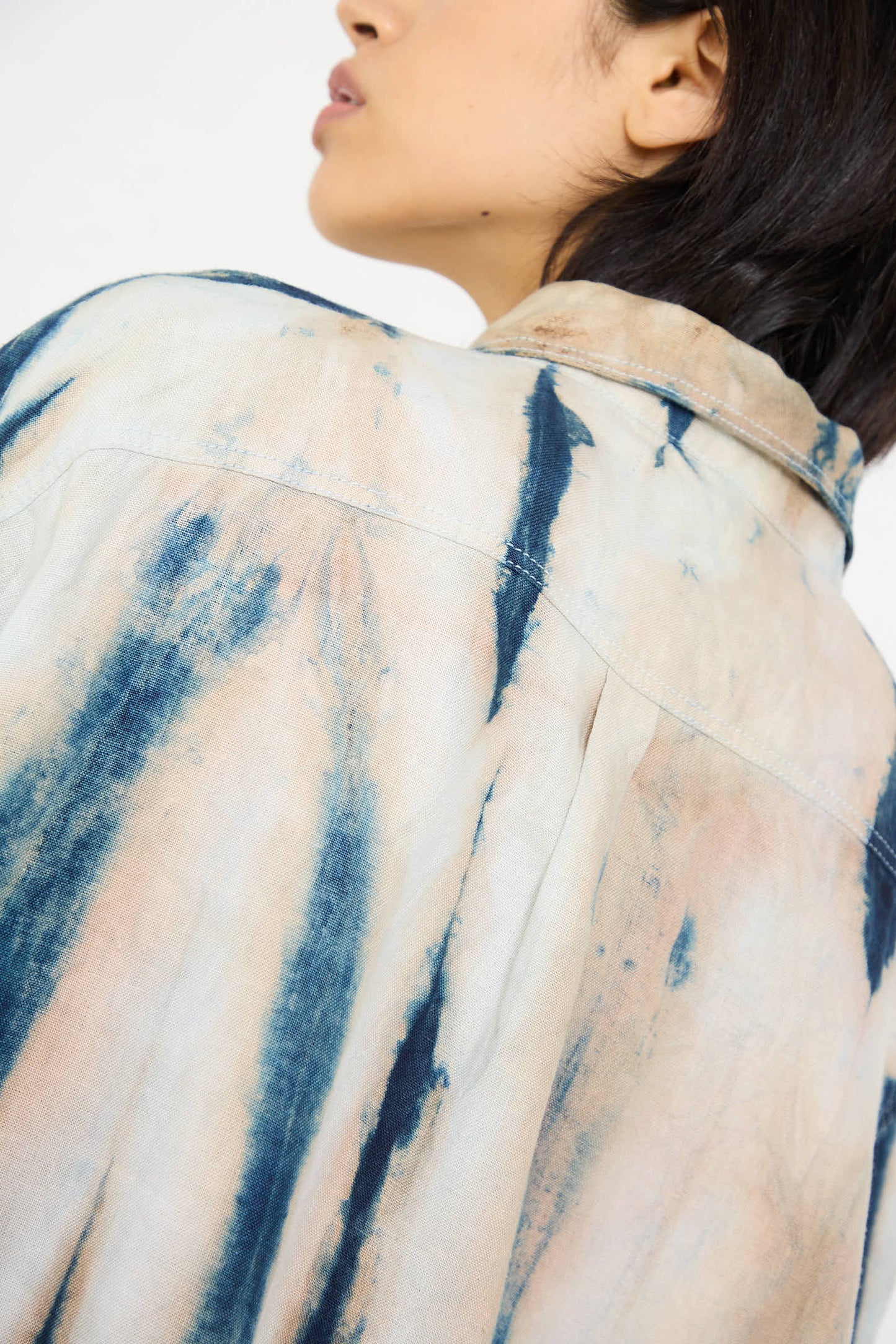 A person with short hair is shown from the back, wearing the Shibori Hemp Overshirt in Faded Indigo by Luna Del Pinal, featuring a light-colored denim fabric with blue tie-dye patterns and a distinctive faded indigo stripe.