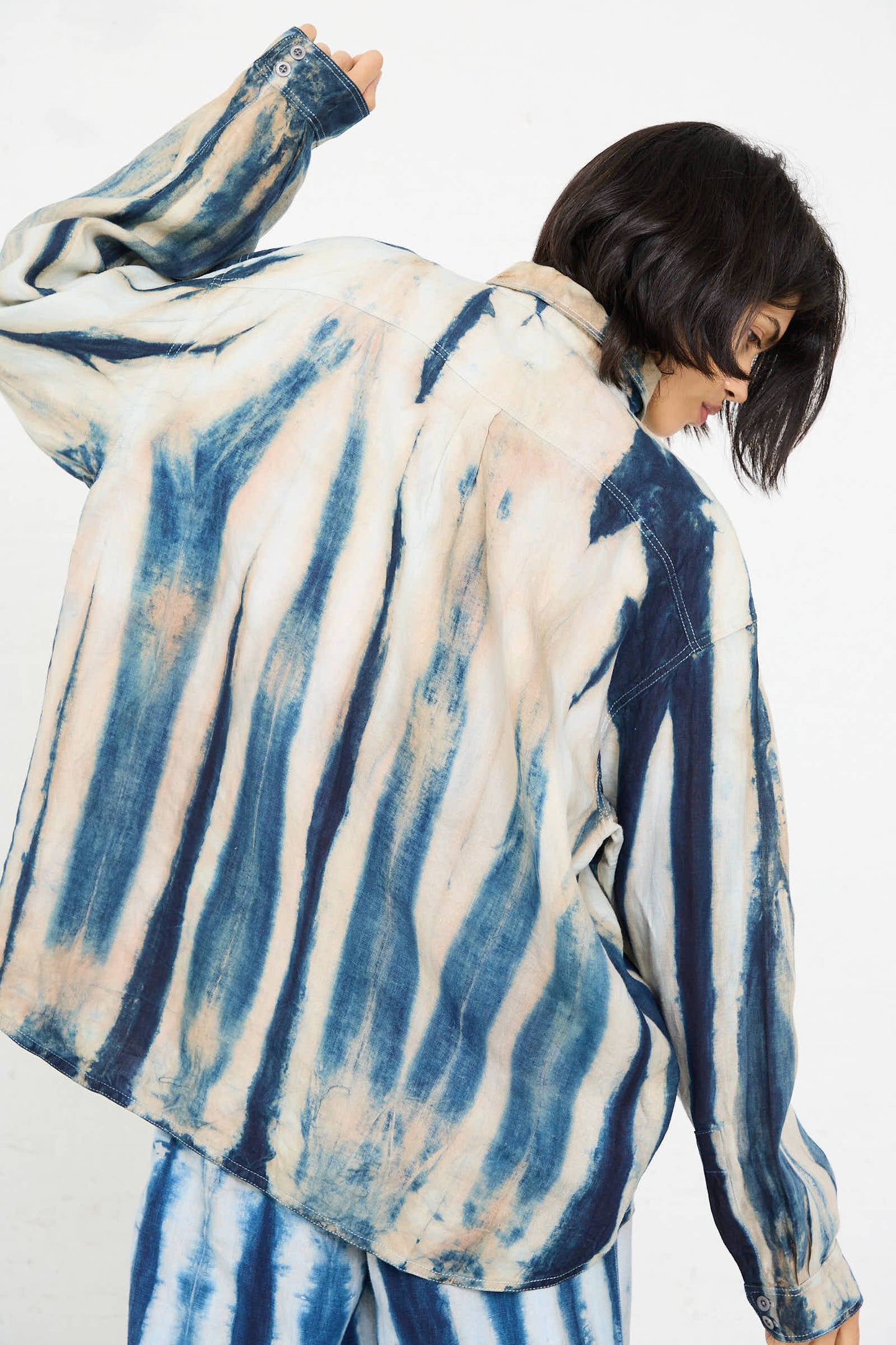 Person with short dark hair wearing the Shibori Hemp Overshirt in Faded Indigo by Luna Del Pinal, made from sustainably grown hemp, along with coordinating pants, viewed from the back with an arm raised.