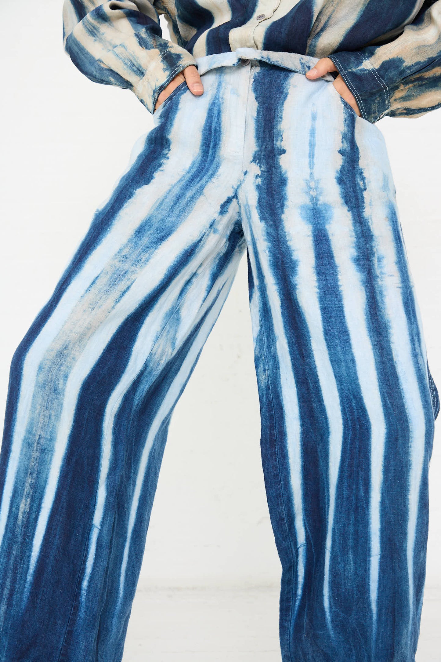 Close-up of a person wearing the Shibori Hemp Wide Leg Trouser in Faded Indigo by Luna Del Pinal, featuring blue and white tie-dye with hands tucked into pockets. The ensemble is completed with a matching top crafted from sustainably grown hemp.