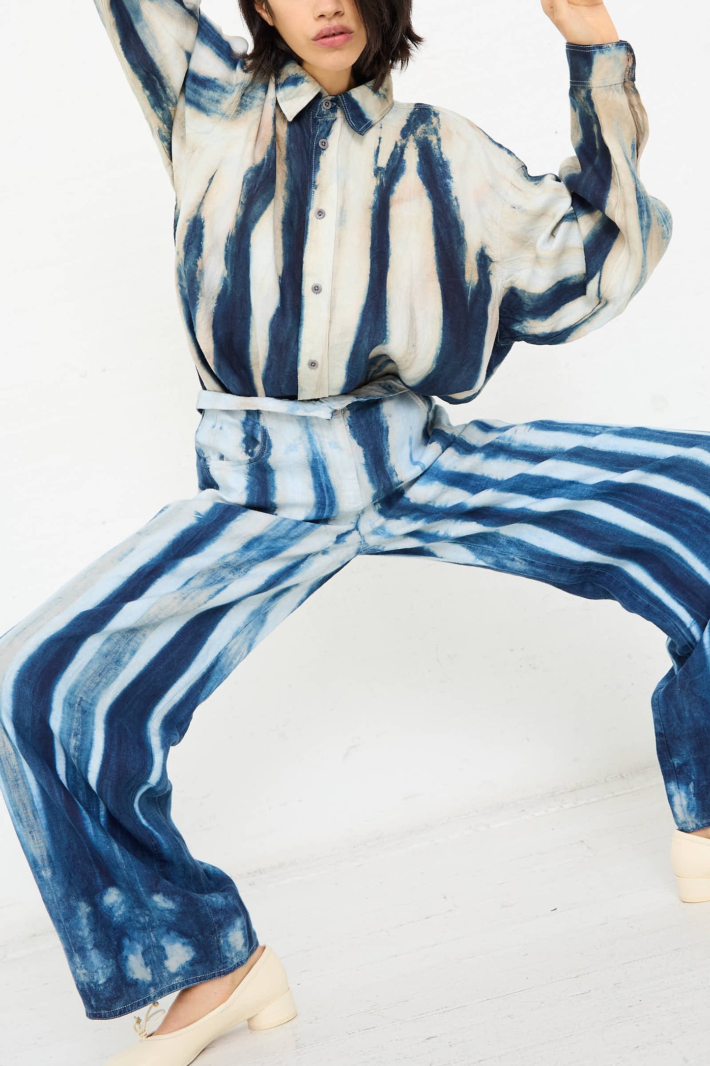 A person donning a matching tie-dye outfit featuring the Luna Del Pinal Shibori Hemp Wide Leg Trouser in Faded Indigo, characterized by blue and white vertical stripes, is posing with knees bent and arms extended against a plain white background.