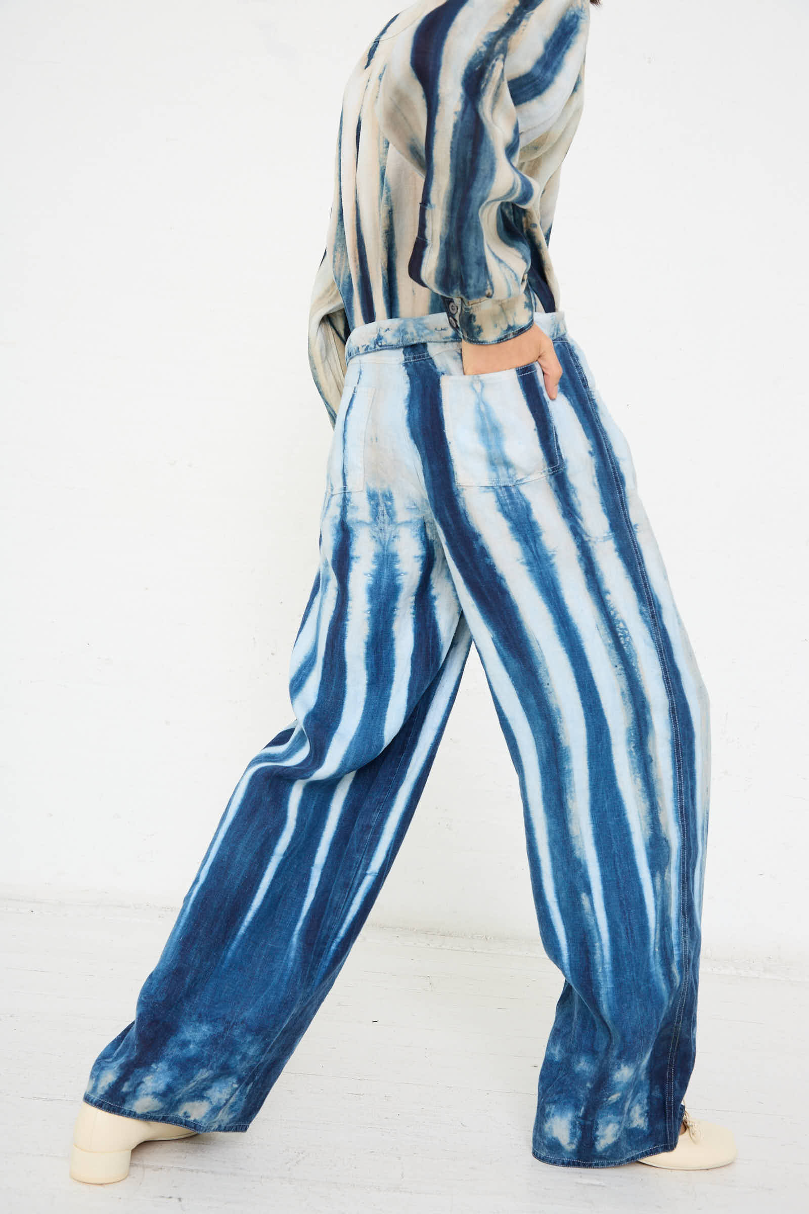 A person dressed in the Shibori Hemp Wide Leg Trouser in Faded Indigo by Luna Del Pinal, with a matching top, is walking with one hand in their pocket against a plain white background.