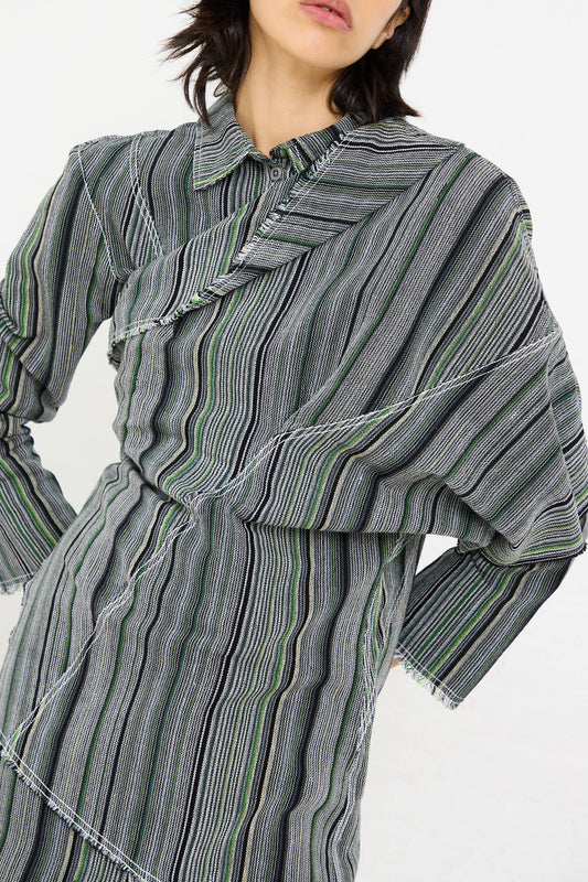 Person wearing the Luna Del Pinal Tela De Raya Loom Weave Asymmetric Top in Humbug Stripe, characterized by long sleeves and shades of green, gray, and black stripes, standing against a white background.