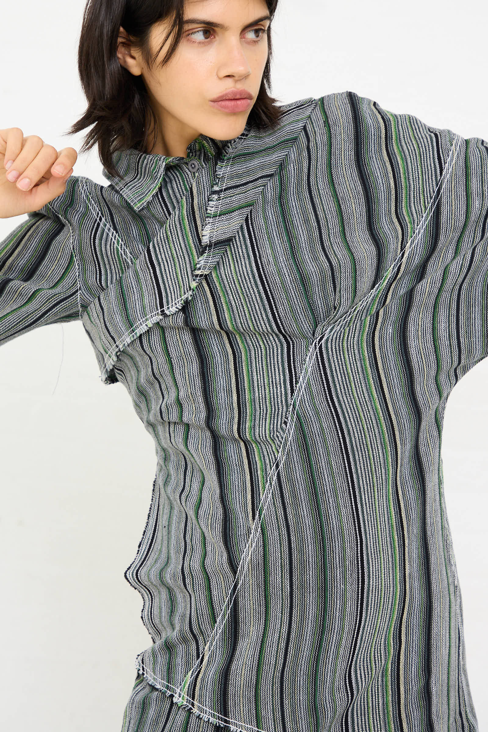 Person wearing the Tela De Raya Loom Weave Asymmetric Top in Humbug Stripe by Luna Del Pinal, featuring a textured, striped cotton design in various shades of gray, green, and black with an asymmetrical neckline, posing against a plain white background.