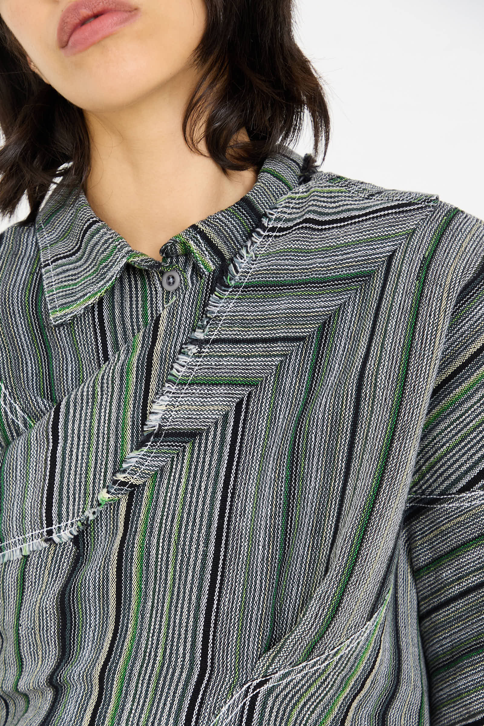 Close-up of a person wearing the Tela De Raya Loom Weave Asymmetric Top in Humbug Stripe by Luna Del Pinal. The top features a mix of green, black, gray, and white lines in a zig-zag pattern with visible buttons and collar.