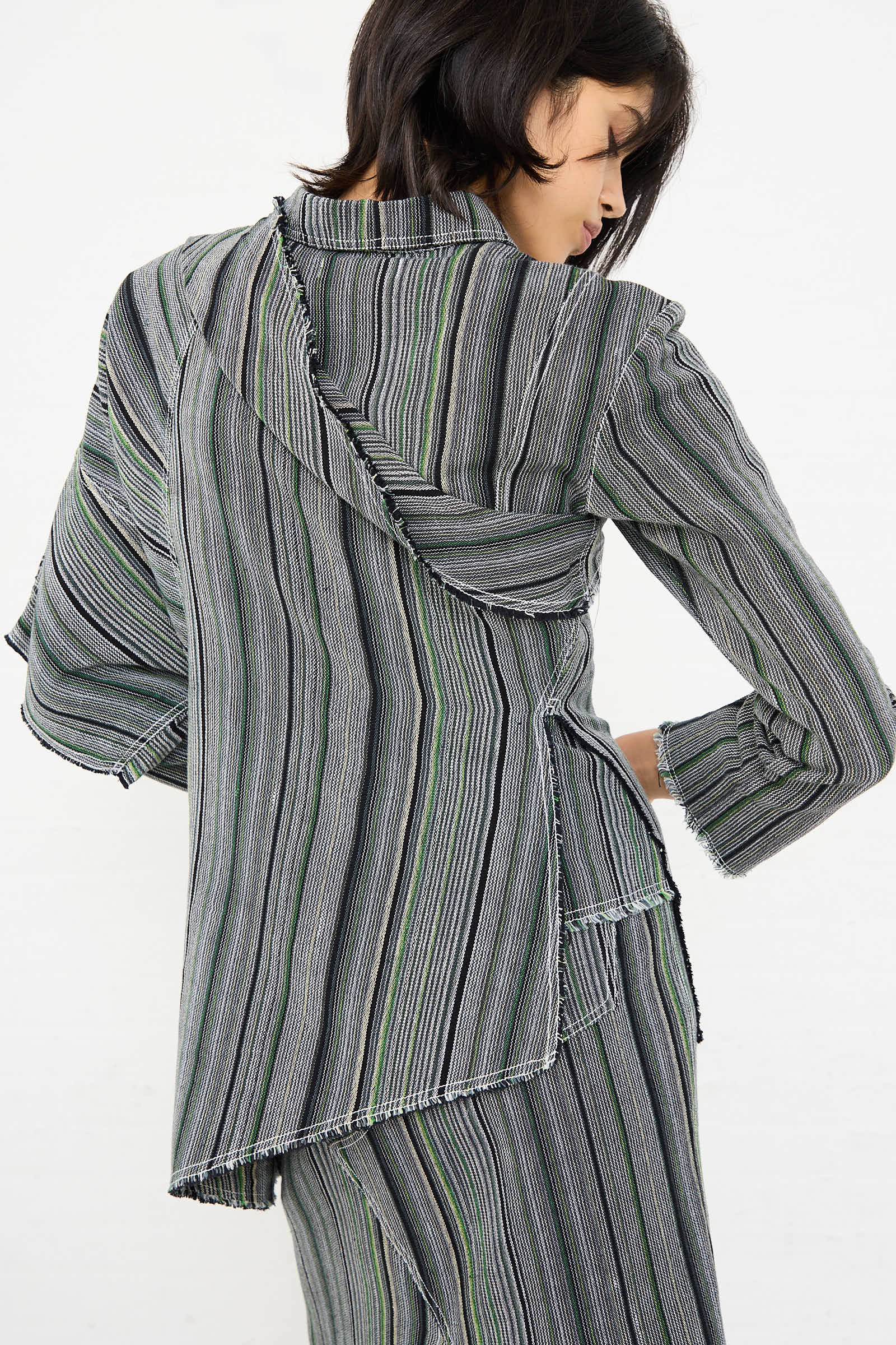 A person is wearing the Tela De Raya Loom Weave Asymmetric Top in Humbug Stripe by Luna Del Pinal. Viewed from the back, this gray and green striped cotton top features an asymmetrical neckline, frayed edges, and a draped, layered effect.