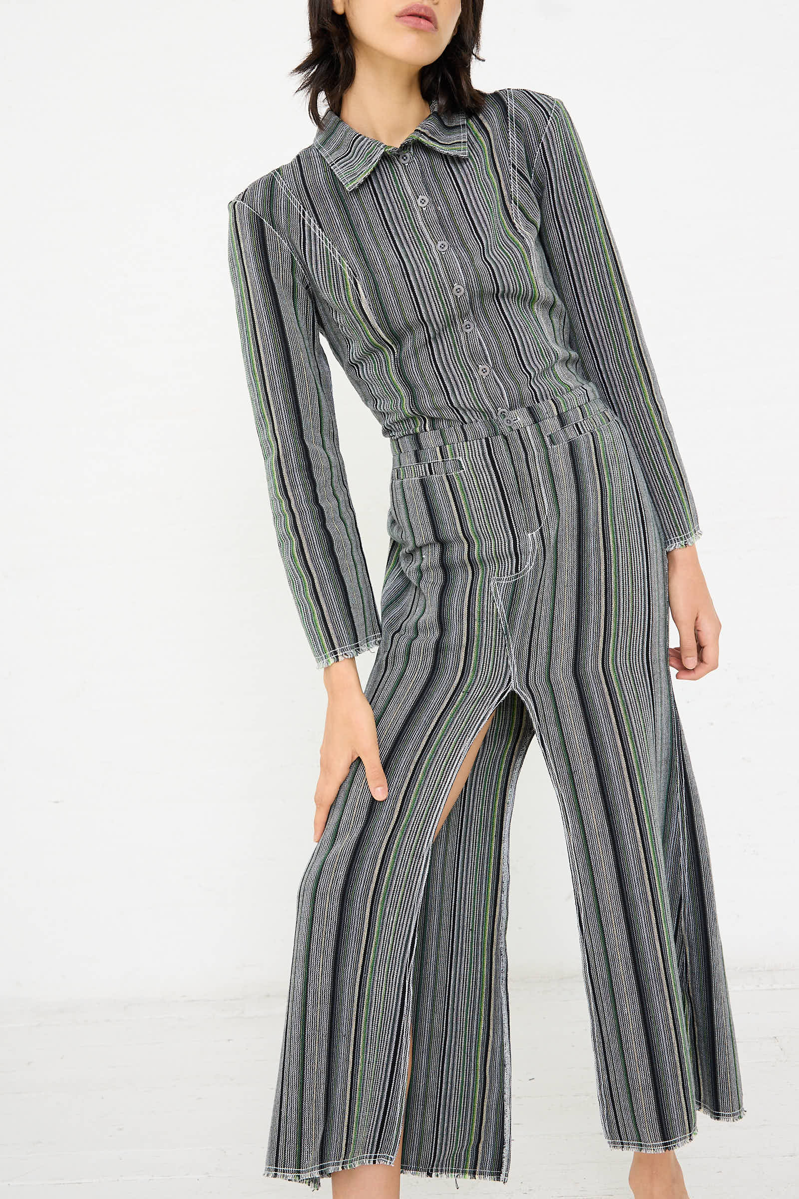 Person wearing Tela De Raya Loom Weave Jean Skirt in Humbug Stripe by Luna Del Pinal, reminiscent of a gray and green pinstriped outfit, consisting of a long-sleeved button-up top and wide-legged pants with a high slit, standing against a white background.