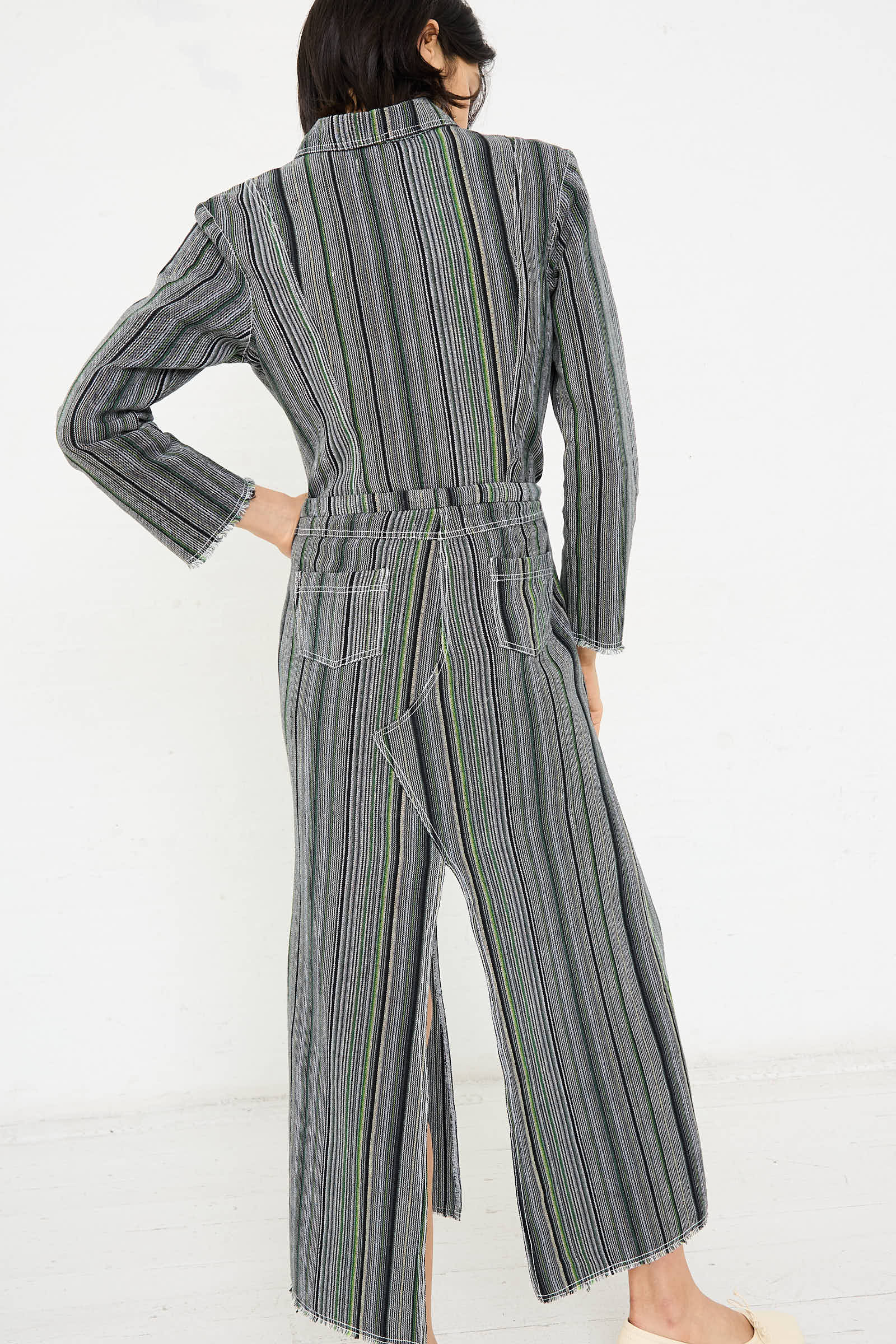 A person is seen from the back, wearing a striped, long-sleeved jumpsuit featuring Humbug Stripe patterns. The jumpsuit showcases a mix of vertical lines in various shades of grey with hints of green and blue.