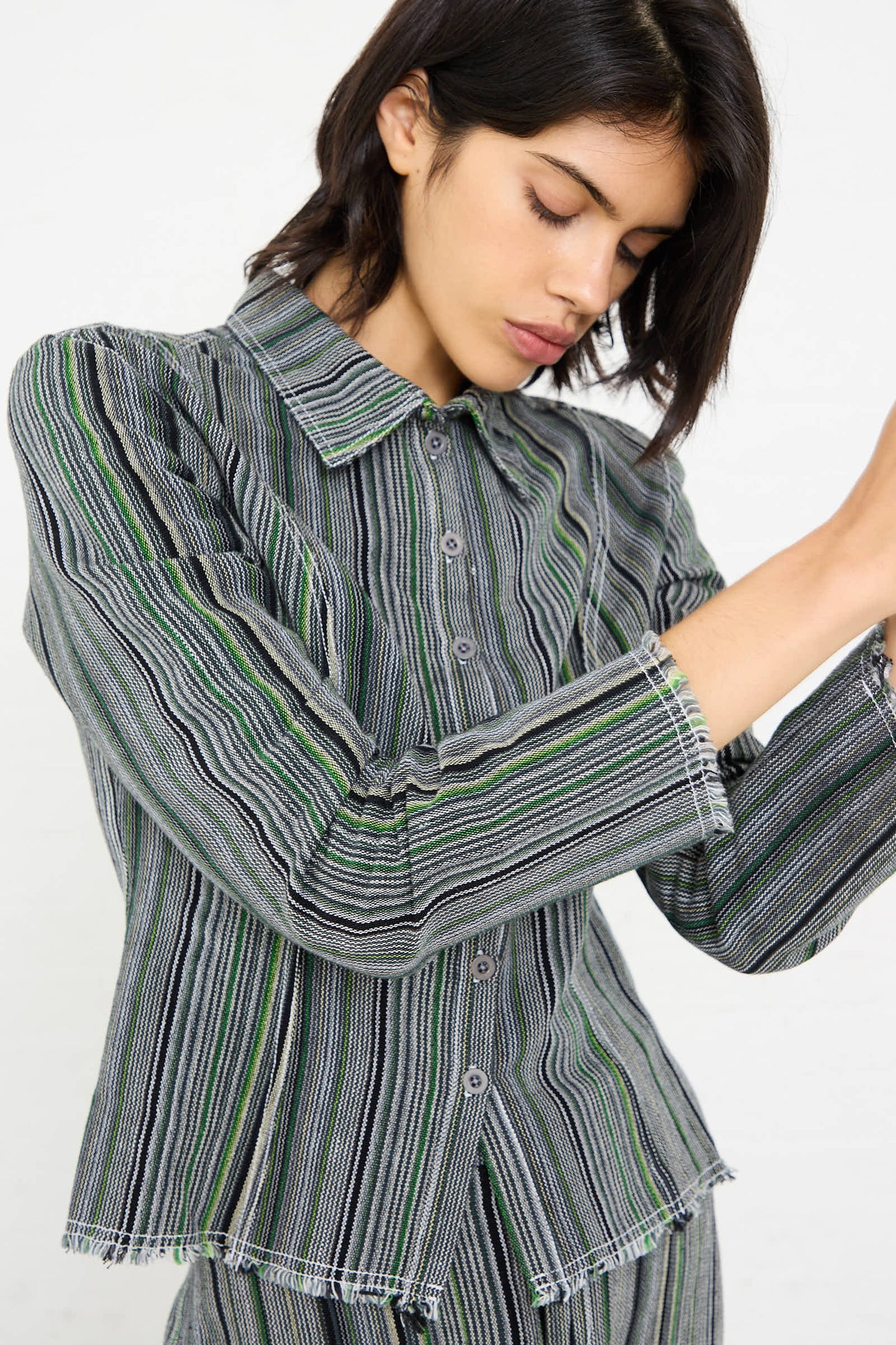 A person with short dark hair, wearing the Tela De Raya Loom Weave Pin Shirt in Humbug Stripe by Luna Del Pinal, poses with their head tilted down and arms raised slightly.