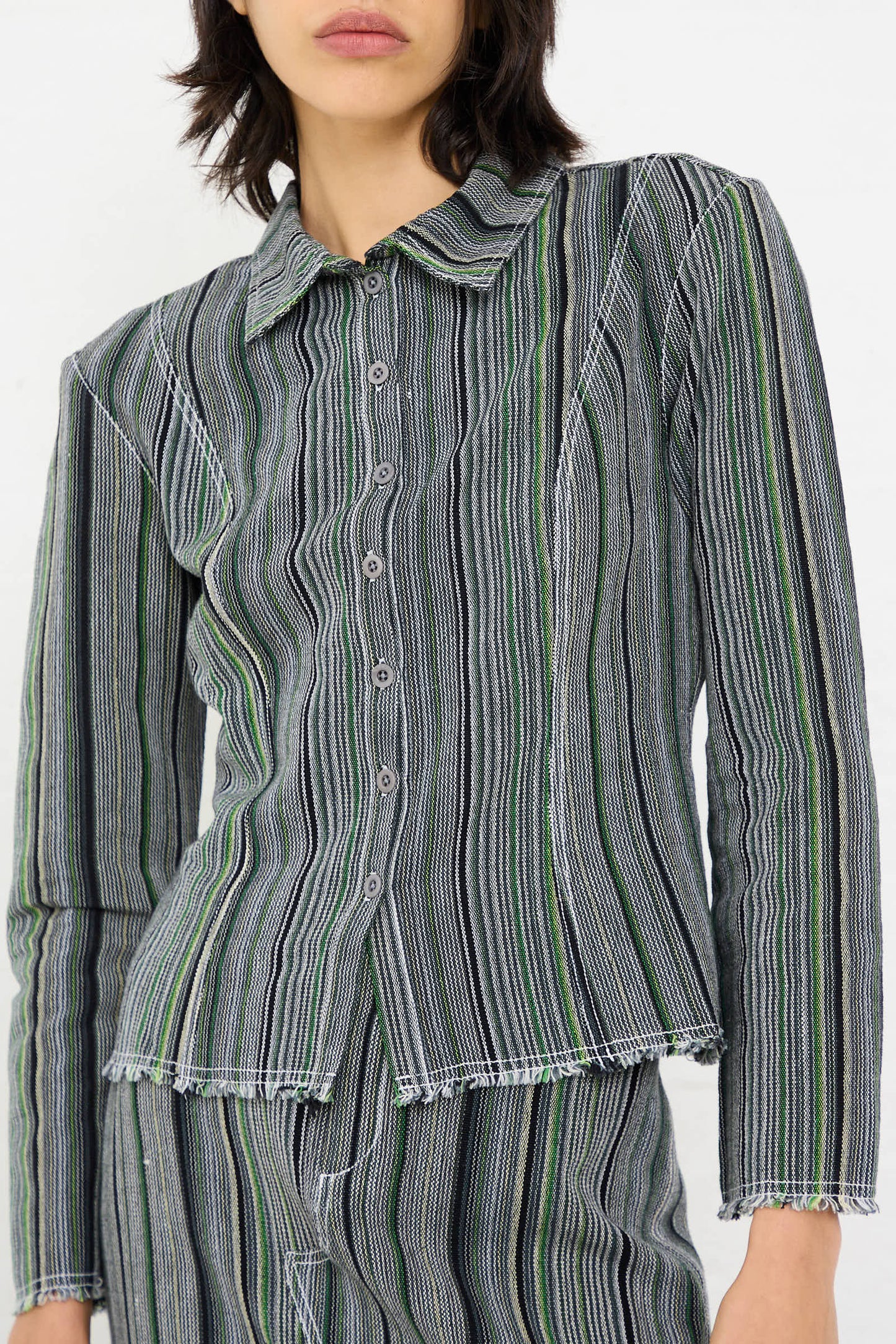 A person is dressed in the Luna Del Pinal Tela De Raya Loom Weave Pin Shirt in Humbug Stripe, a long sleeve button-up featuring black, white, and green stripes with frayed hems. They have paired it with matching pants that share the same subtle grey stripes. Their head is slightly turned, revealing their short, dark hair.