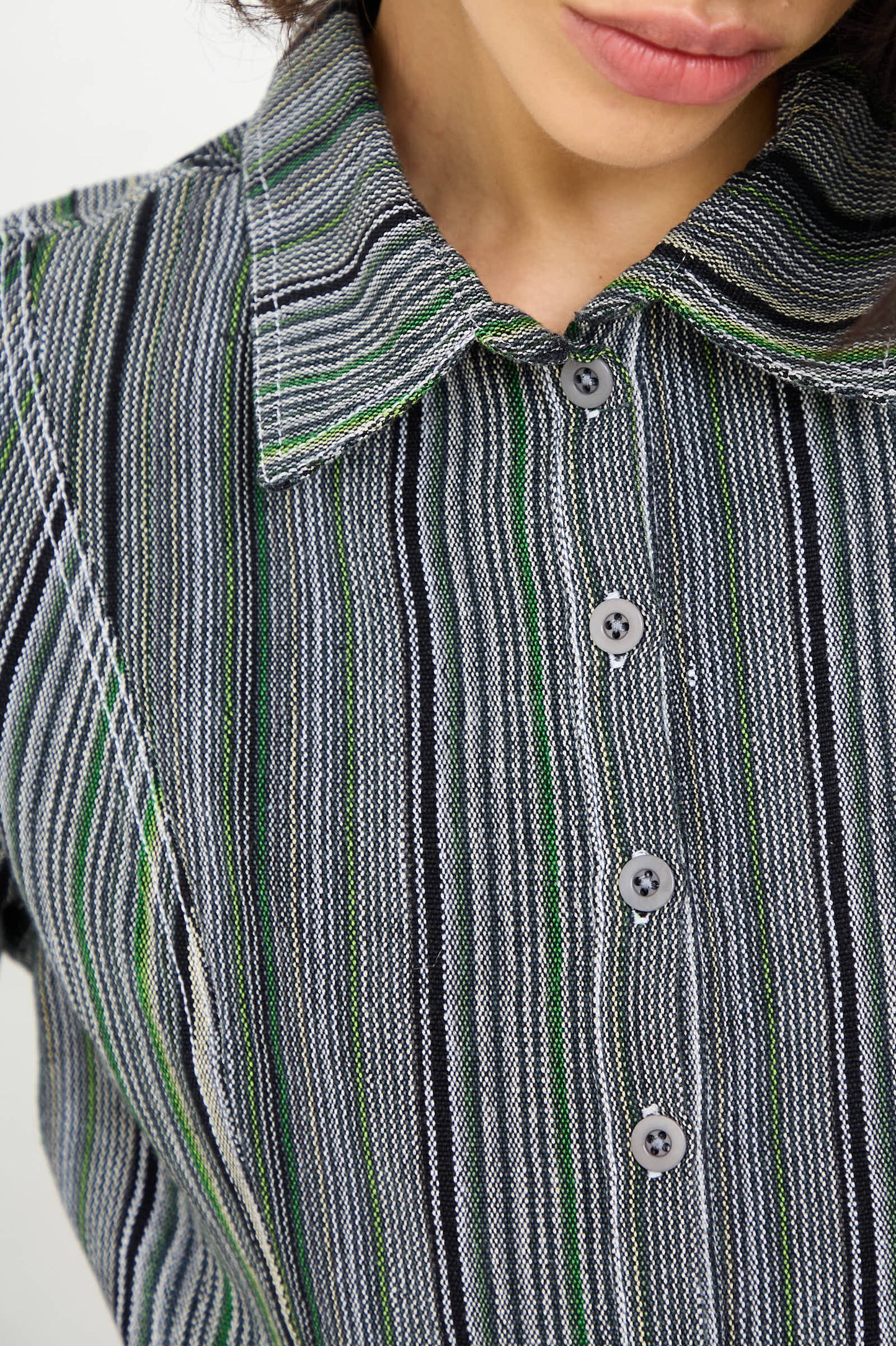 Close-up of a person wearing the Tela De Raya Loom Weave Pin Shirt in Humbug Stripe by Luna Del Pinal, which features a buttoned-up, long sleeve design with a collar. The shirt displays an intricate pattern of black, white, green, and grey vertical stripes. The person's face is partially visible in the cropped silhouette.