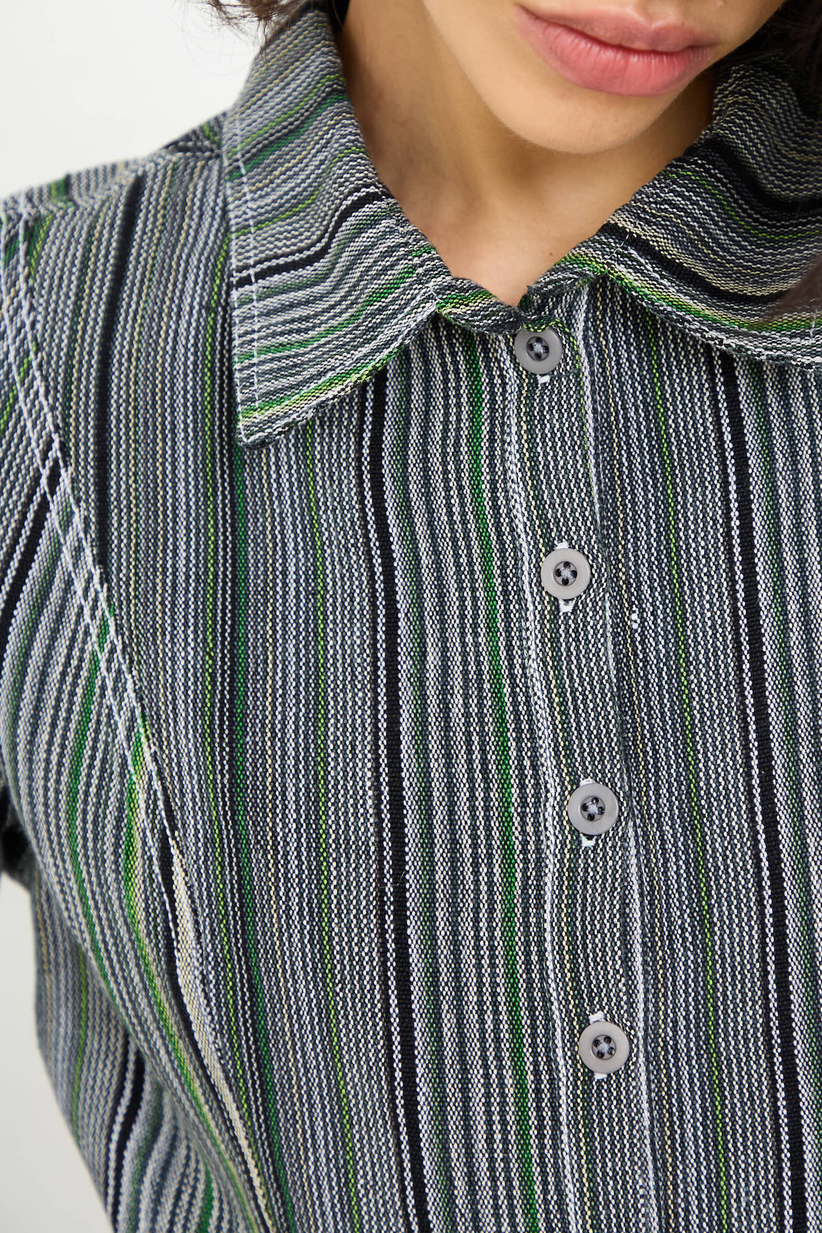 Close-up of a person wearing the Tela De Raya Loom Weave Pin Shirt in Humbug Stripe by Luna Del Pinal, which features a buttoned-up, long sleeve design with a collar. The shirt displays an intricate pattern of black, white, green, and grey vertical stripes. The person's face is partially visible in the cropped silhouette.