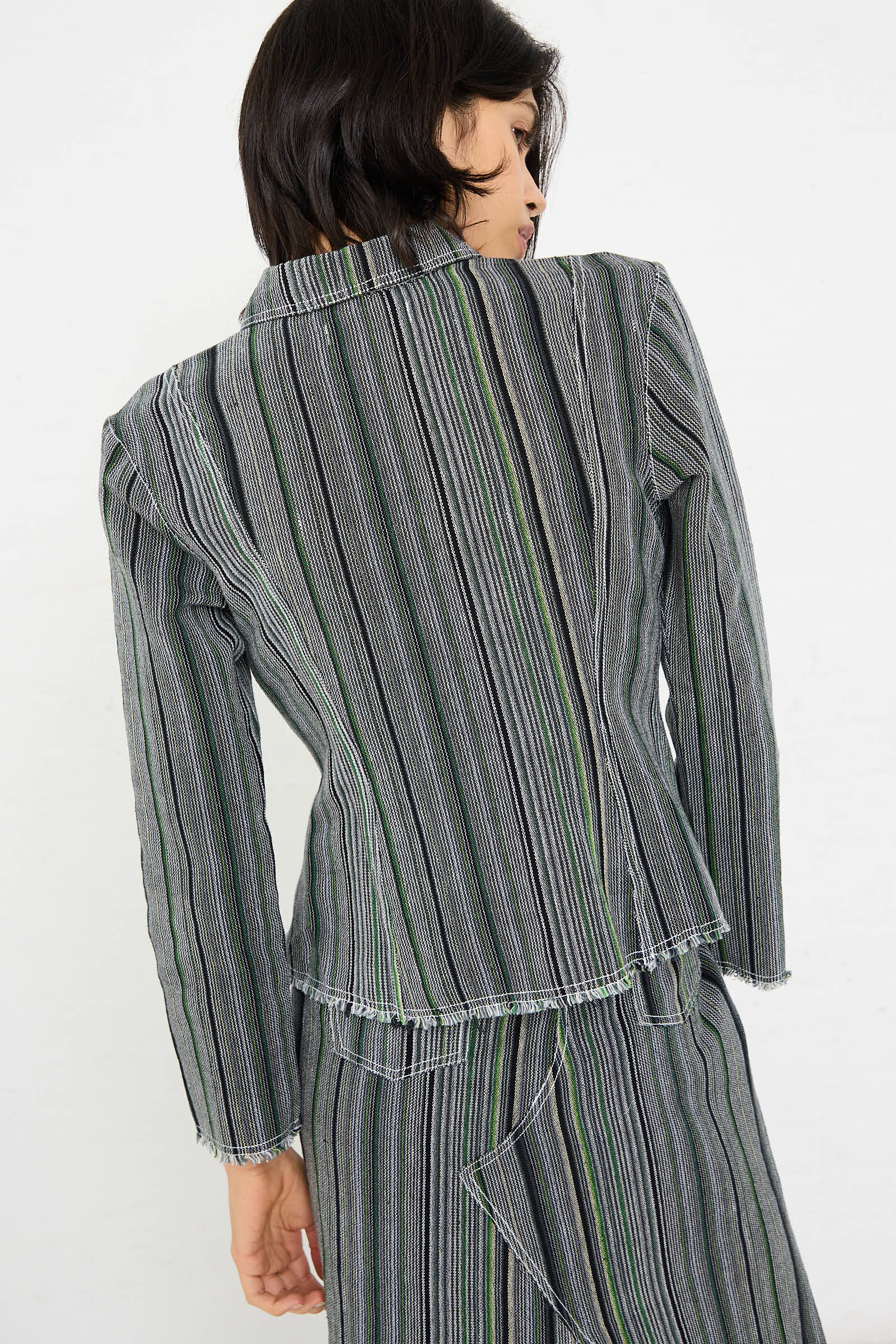 A woman wearing the Tela De Raya Loom Weave Pin Shirt in Humbug Stripe by Luna Del Pinal with vertically striped, long sleeves and frayed edges, viewed from the back, along with a matching grey striped skirt visible.