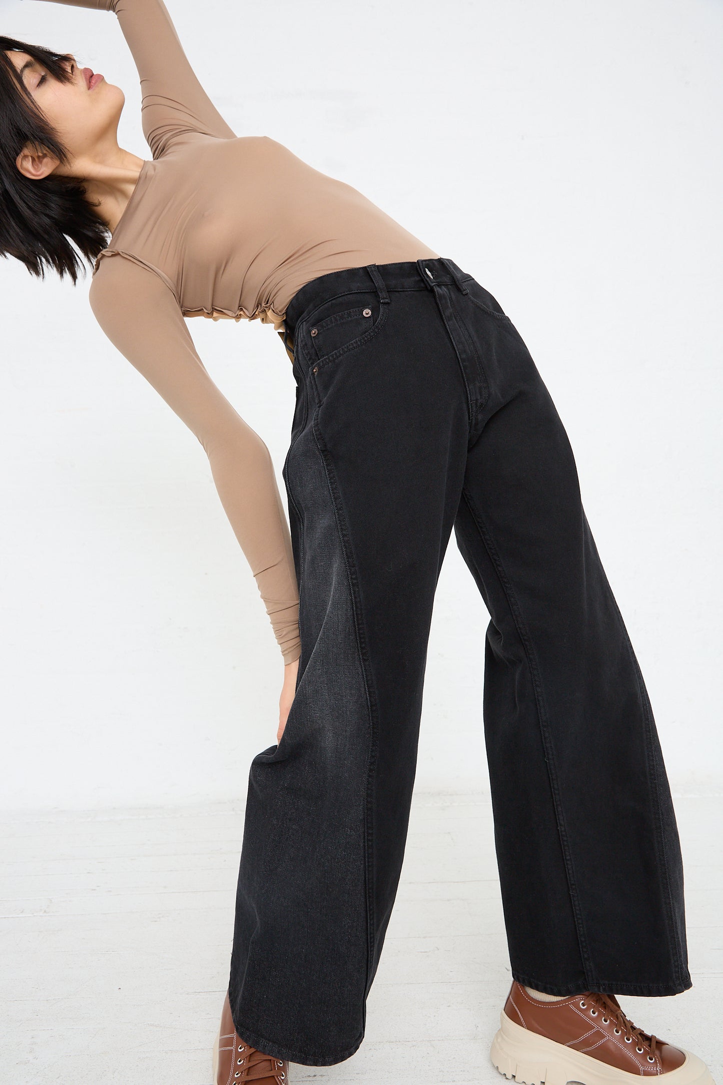 Woman in MM6 wide-leg black denim jeans and a beige top stretching to one side against a white background.
