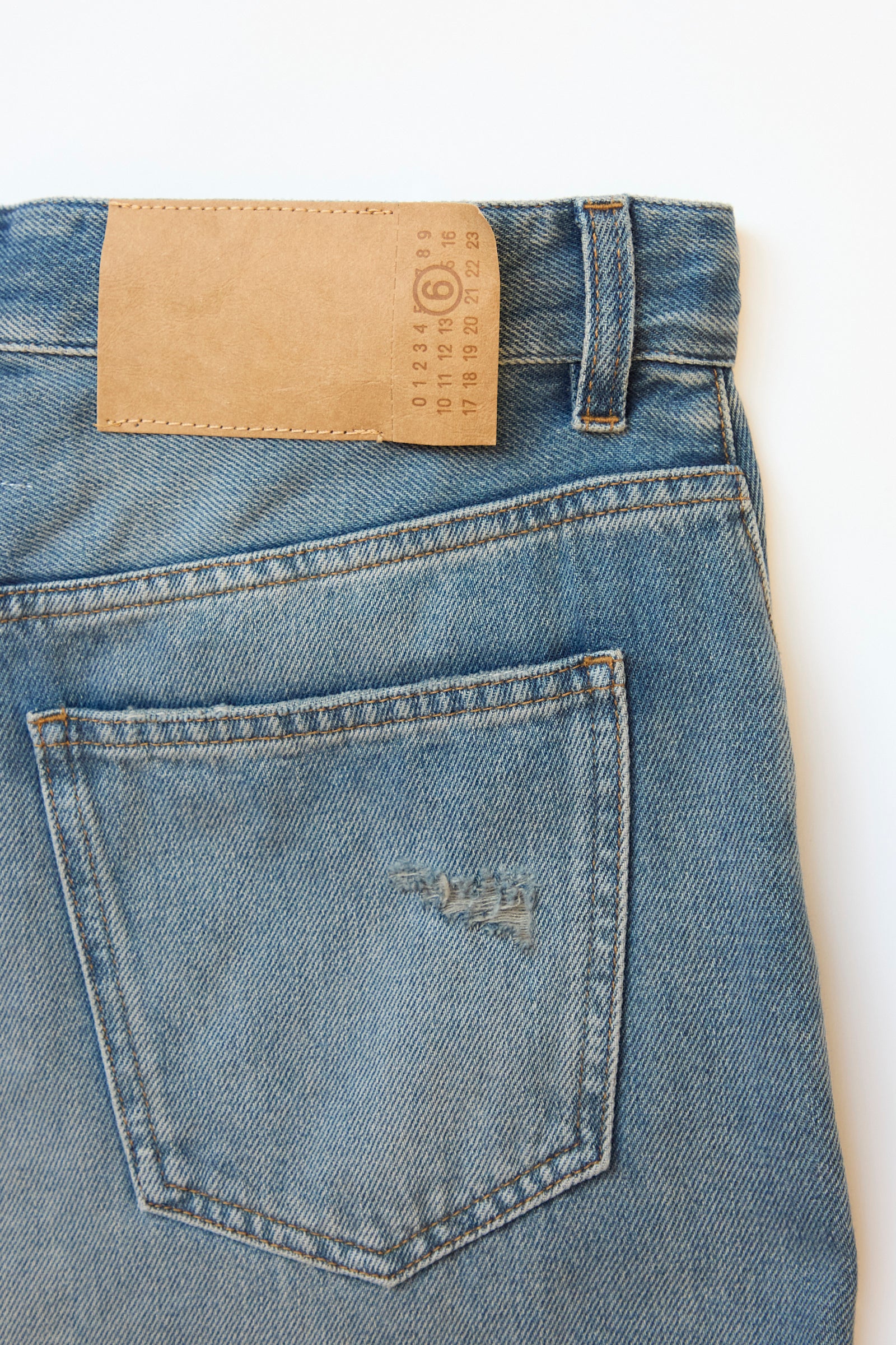 A close-up shows the back pocket of MM6's 5 Pocket Pant in Grey Blue, highlighting a leather patch with numerical details and a stitched logo.