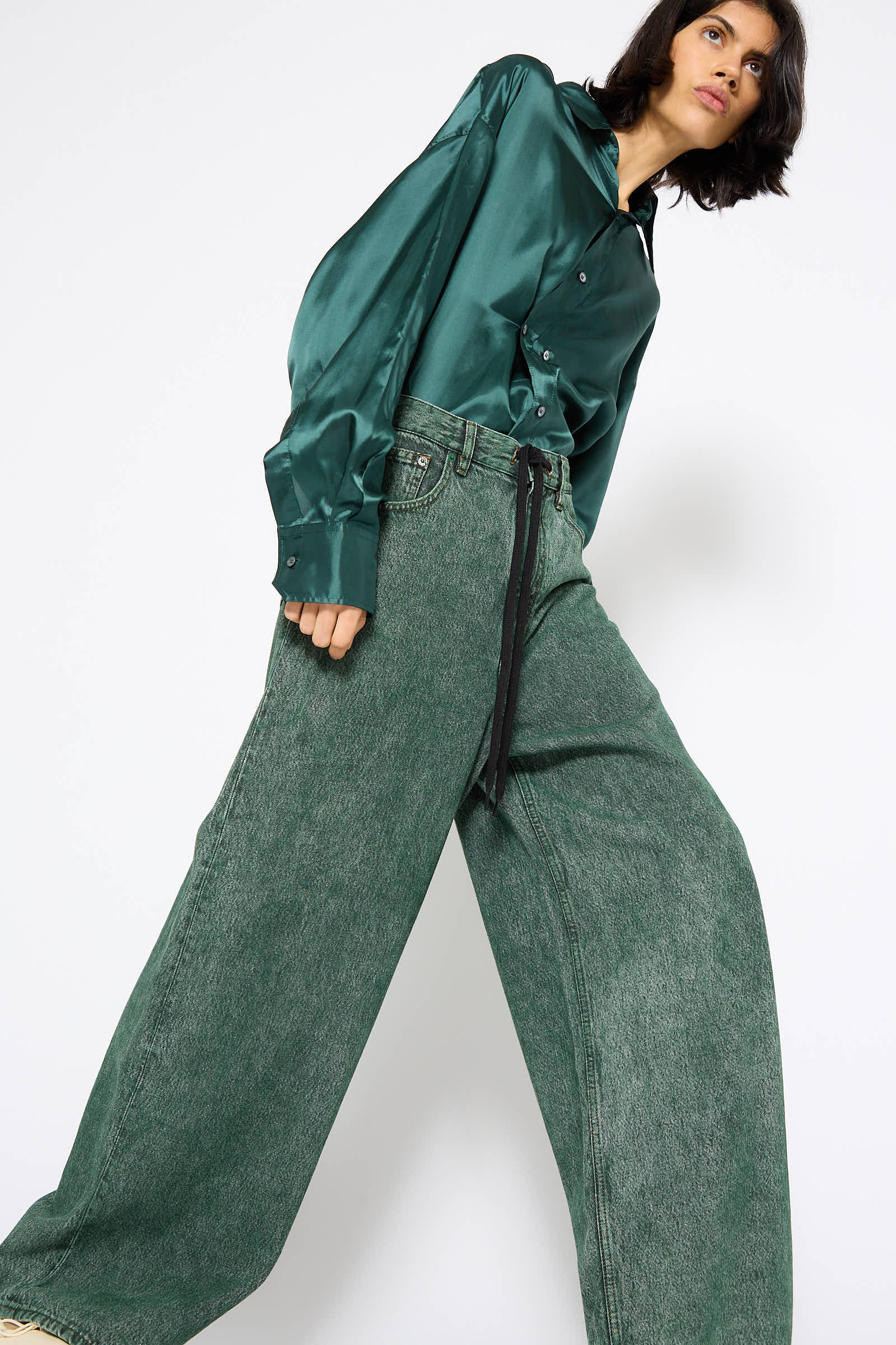 A person stands wearing a dark green satin shirt and MM6's 5 Pockets Denim Pant in Green with a black belt, against a white background.