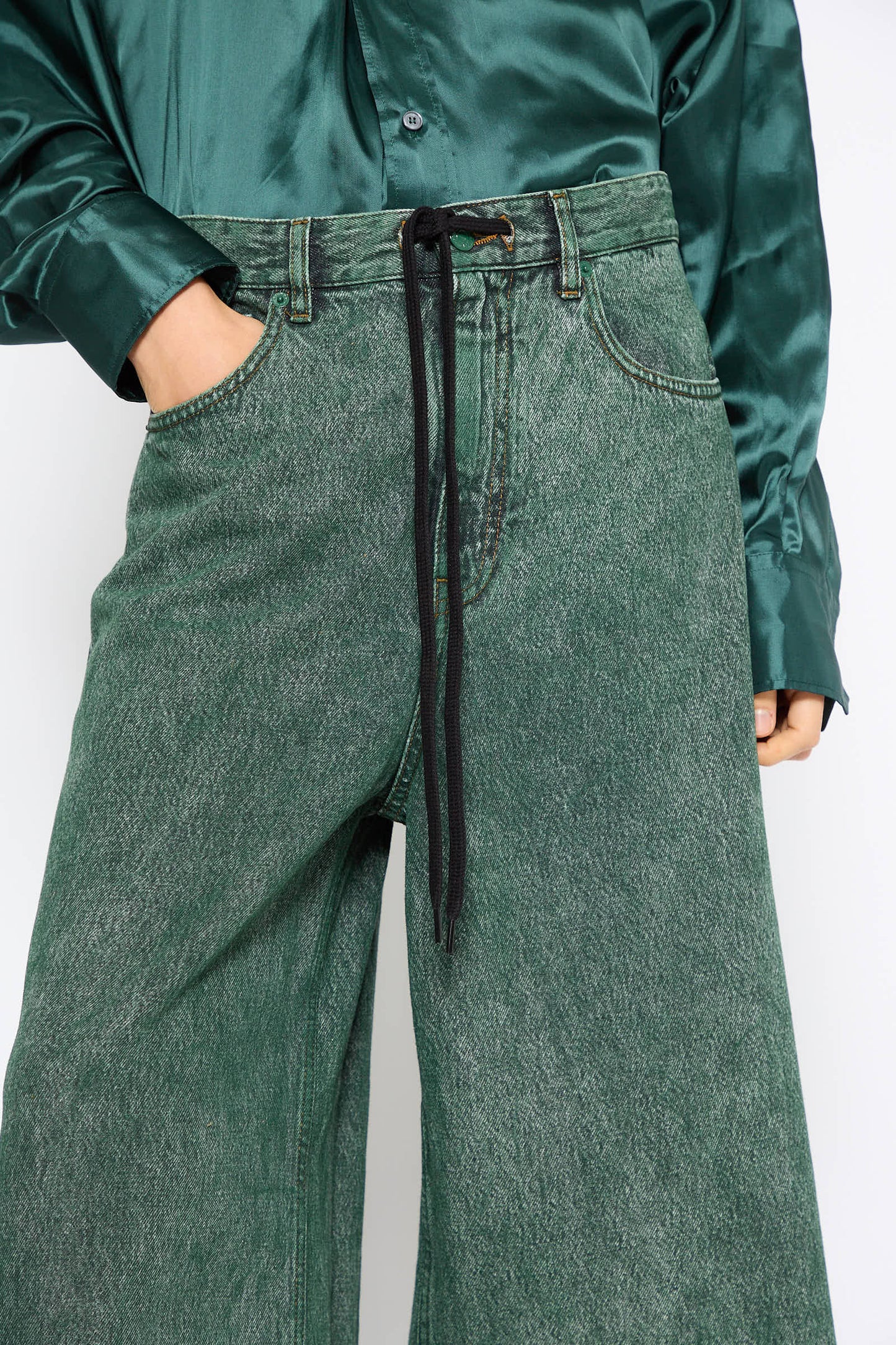 A person wearing a dark green satin shirt and MM6 5 Pockets Denim Pant in Green with a black drawstring belt stands with one hand in their pocket.