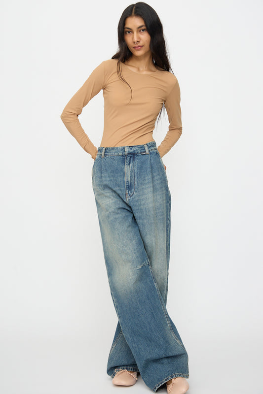 A woman stands against a plain backdrop wearing a beige long-sleeve top, MM6 5 Pocket Pants in Blue with a relaxed wide-leg fit, and nude flats.