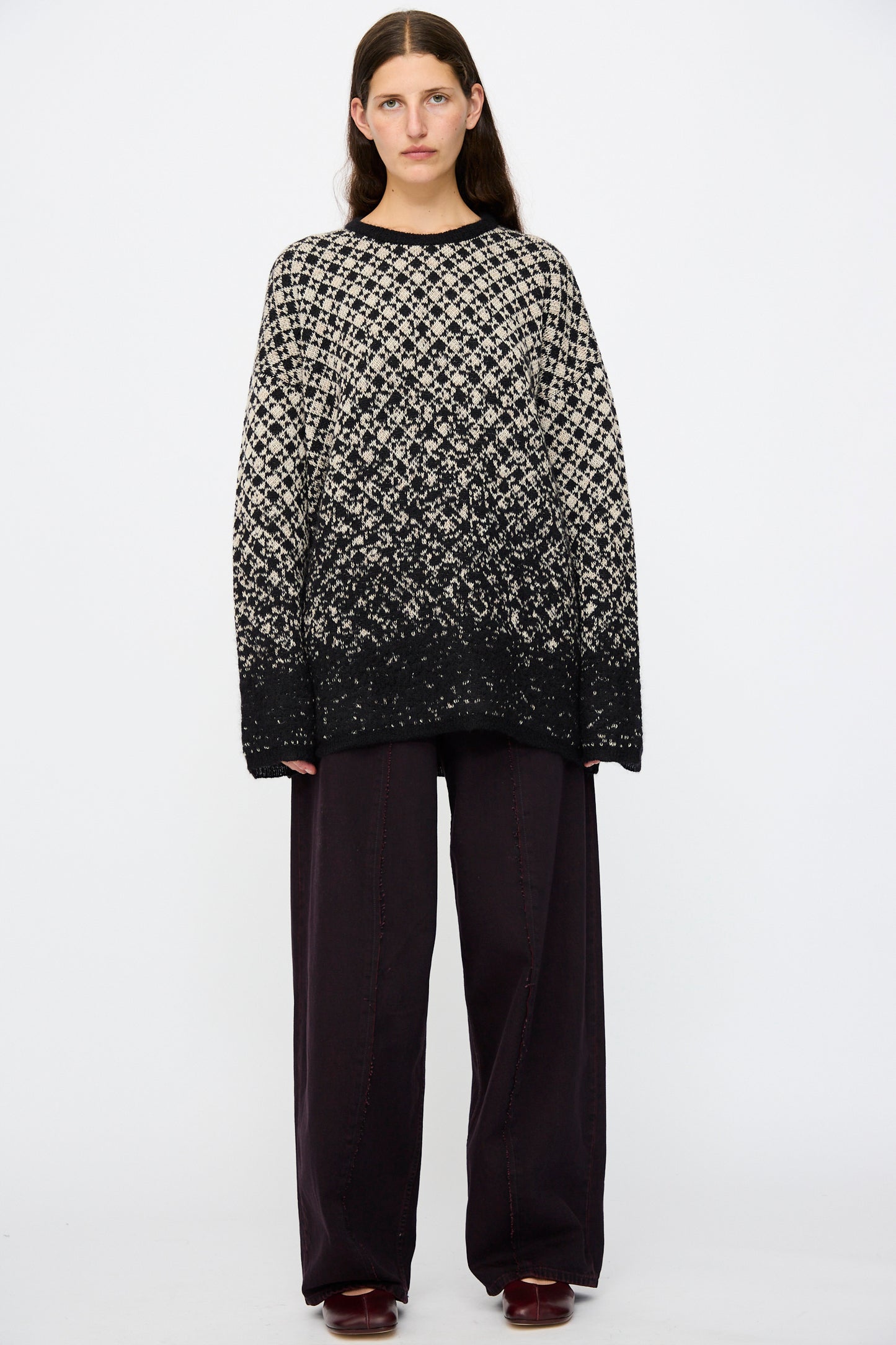 A person stands against a plain background wearing a loose, patterned black-and-white sweater, MM6 5 Pocket Pants in Burgundy, and dark shoes.
