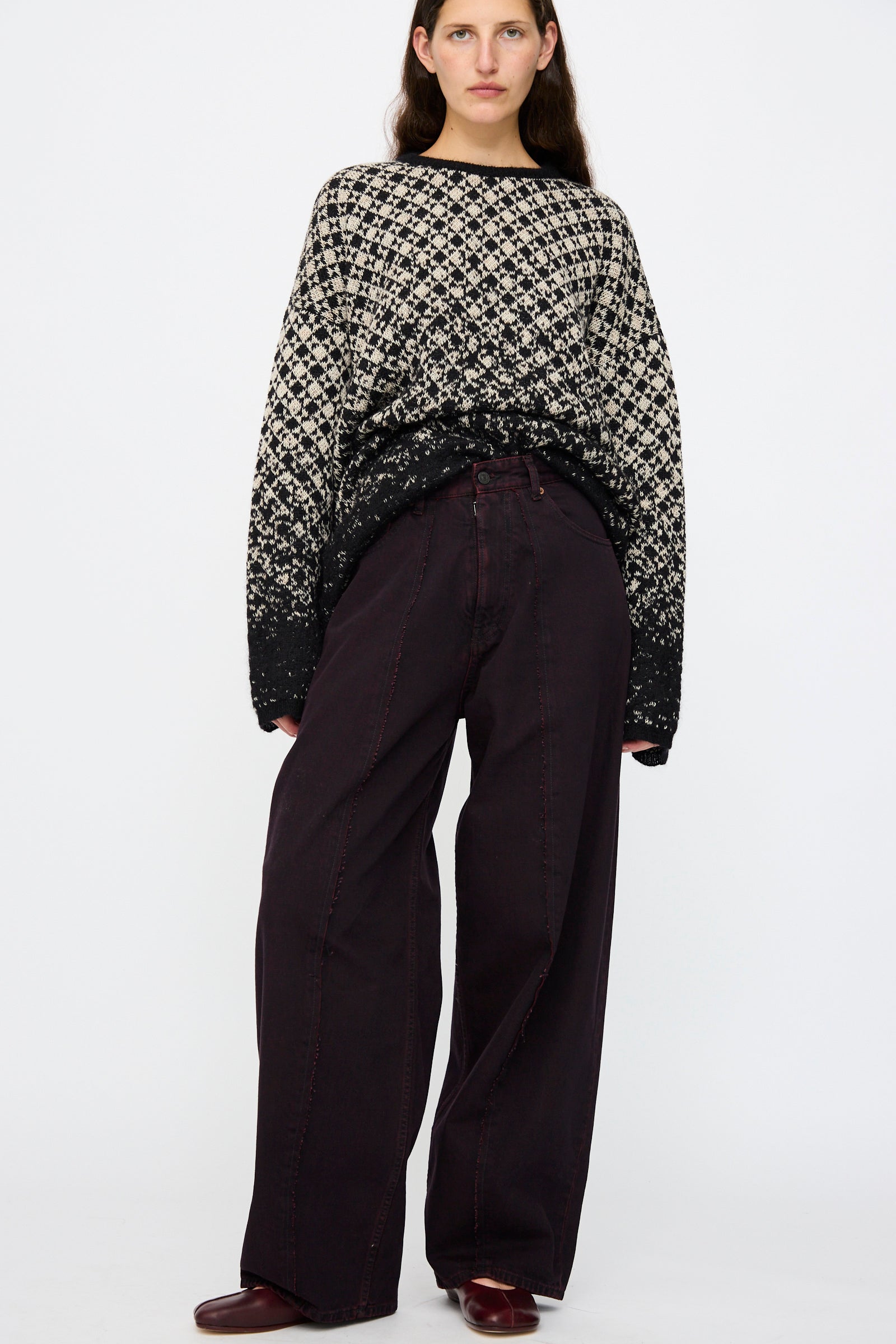 A person stands against a plain background wearing a patterned black and white sweater and MM6 5 Pocket Pants in Burgundy.