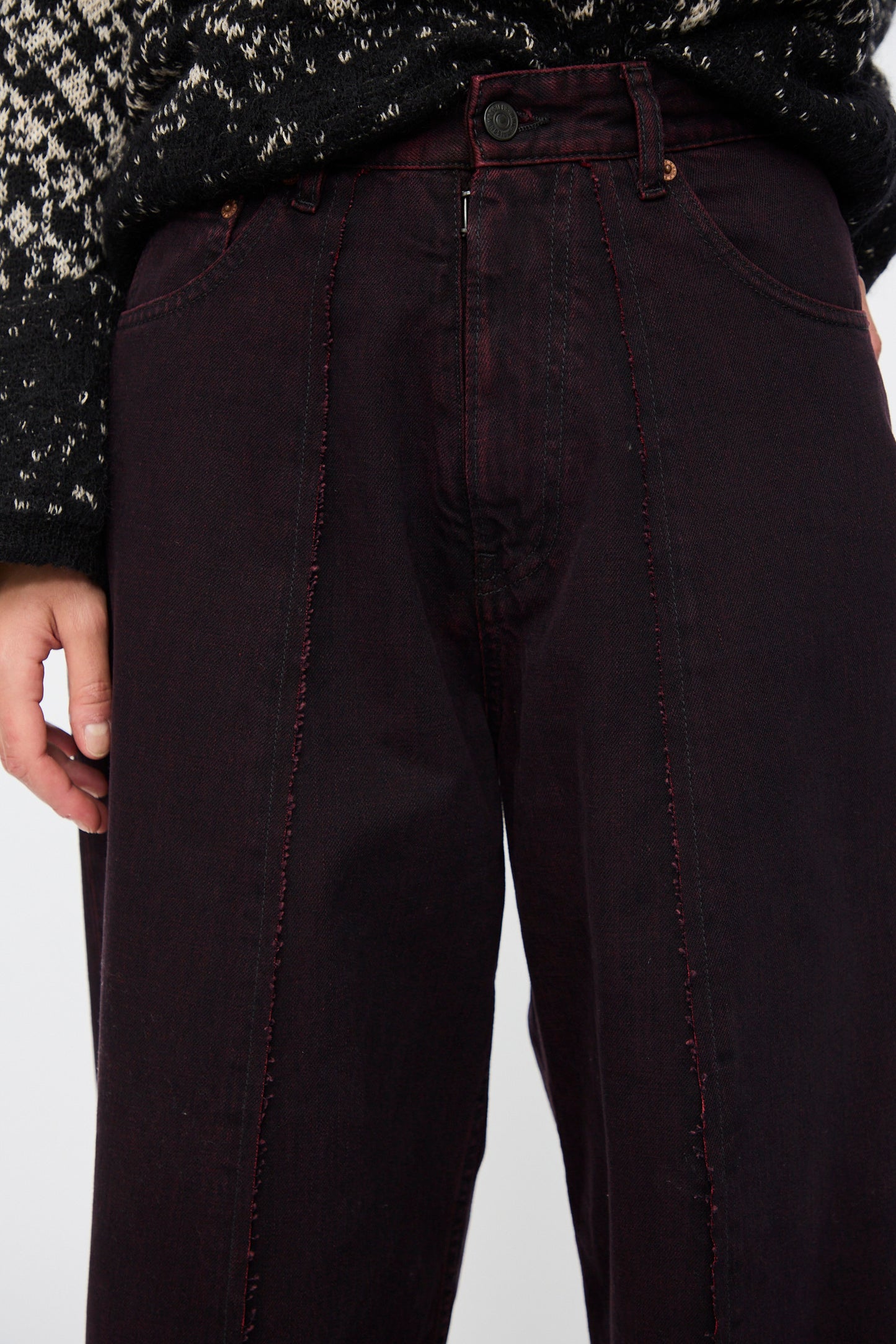 A person wearing MM6’s 5 Pocket Pants in Burgundy—high-waisted, wide leg cotton denim with front seam stitching—paired with a patterned black and white sweater.