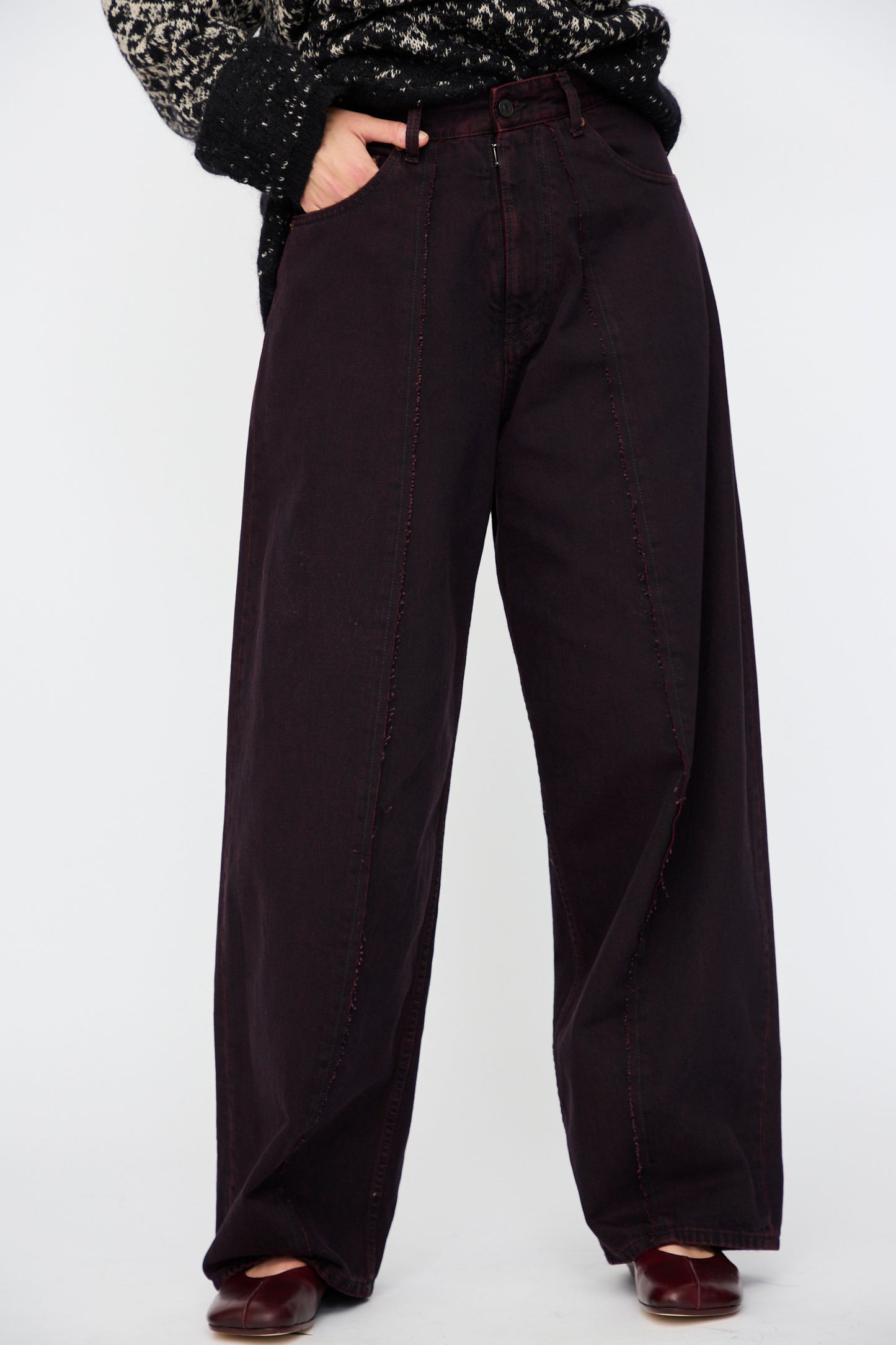 A person wears MM6 5 Pocket Pants in Burgundy, paired with a black and white patterned sweater, and stands against a plain light background.