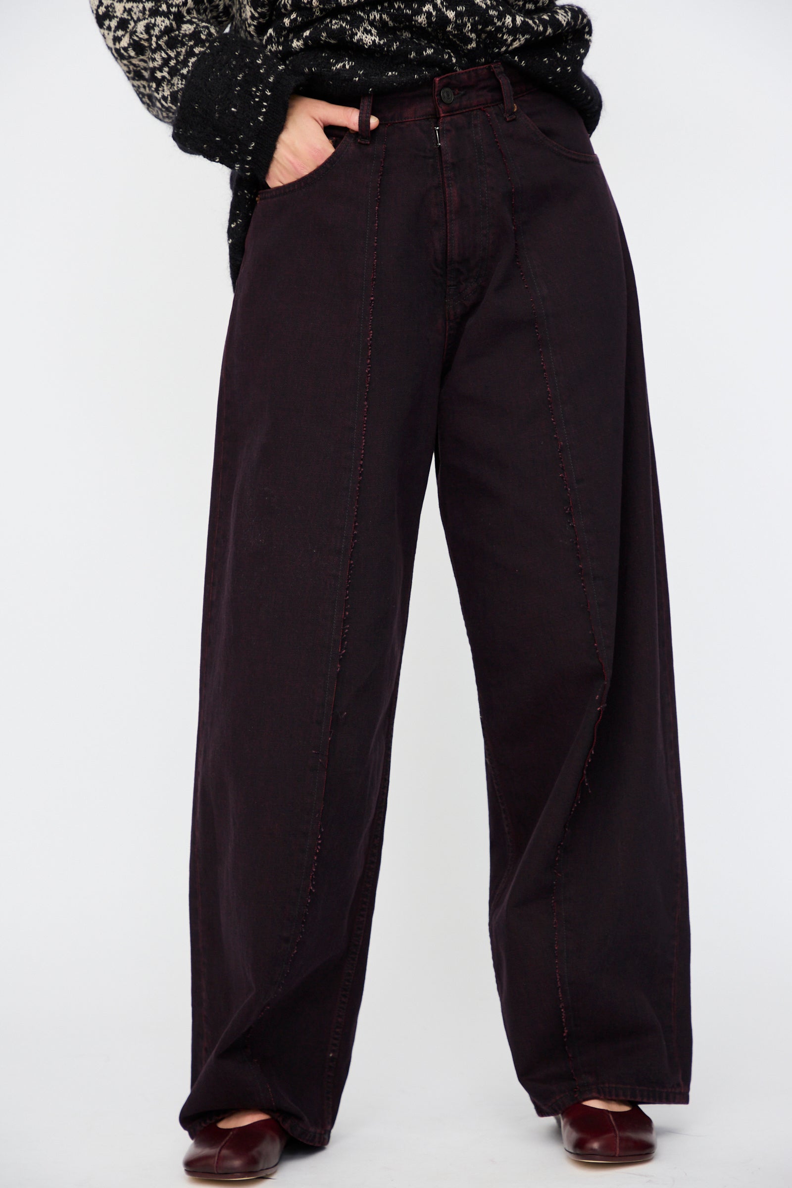 A person wears MM6 5 Pocket Pants in Burgundy, paired with a black and white patterned sweater, and stands against a plain light background.