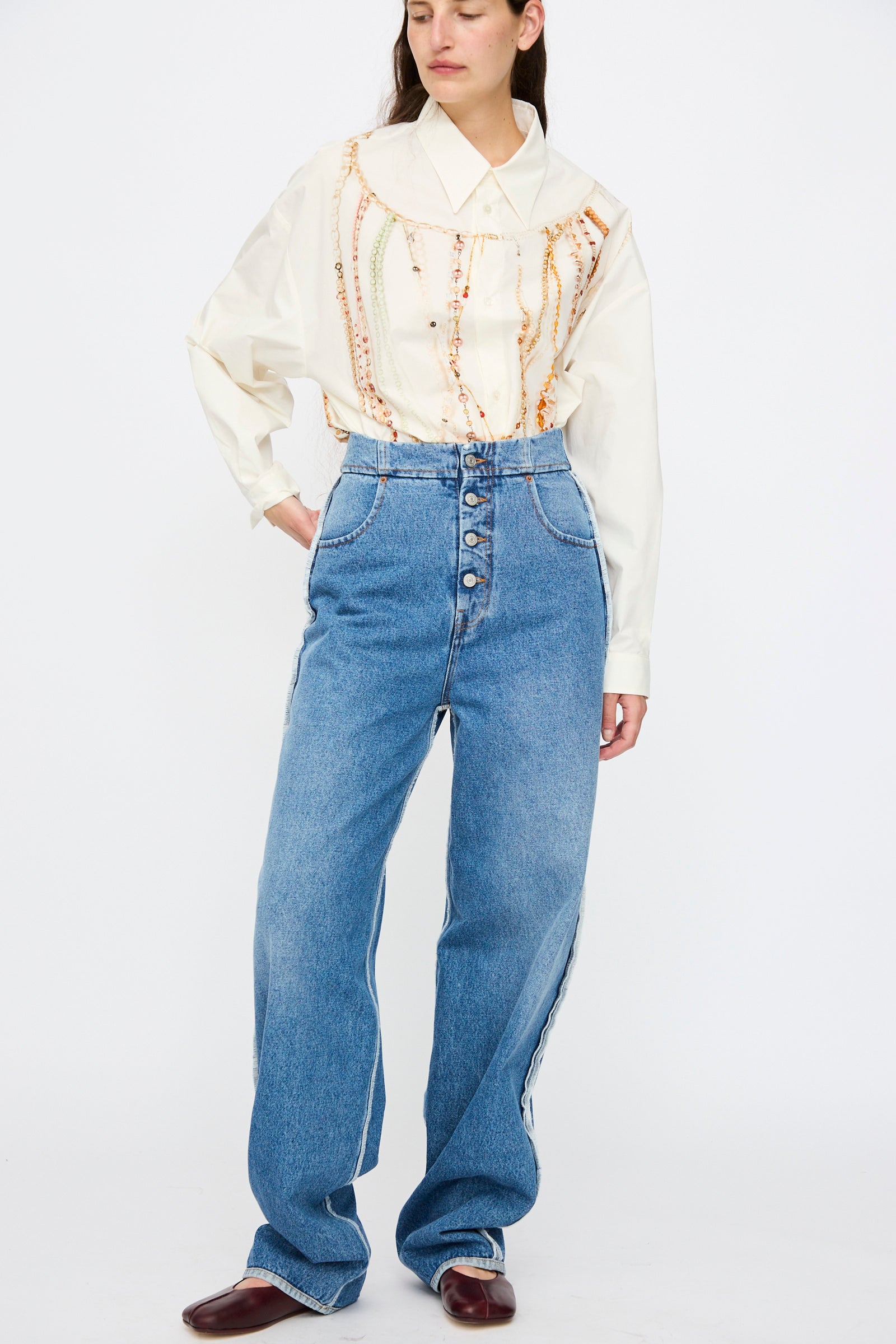 A woman stands against a white background wearing a cream blouse with embroidery, MM6 5 Pocket Pants in Medium Blue with visible buttons, and brown shoes.