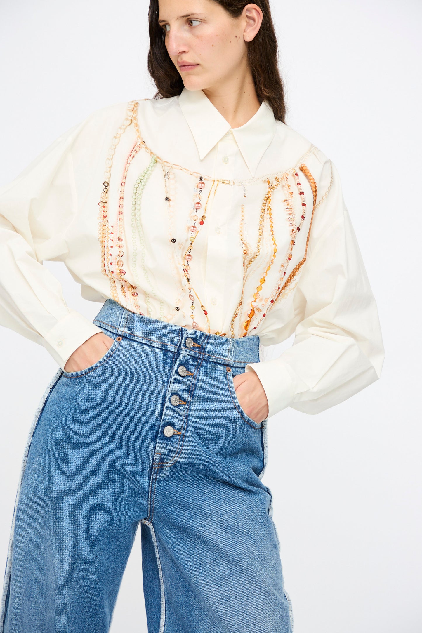 A woman wearing a cream blouse with decorative beading and MM6 5 Pocket Pants in Medium Blue stands against a plain white background.