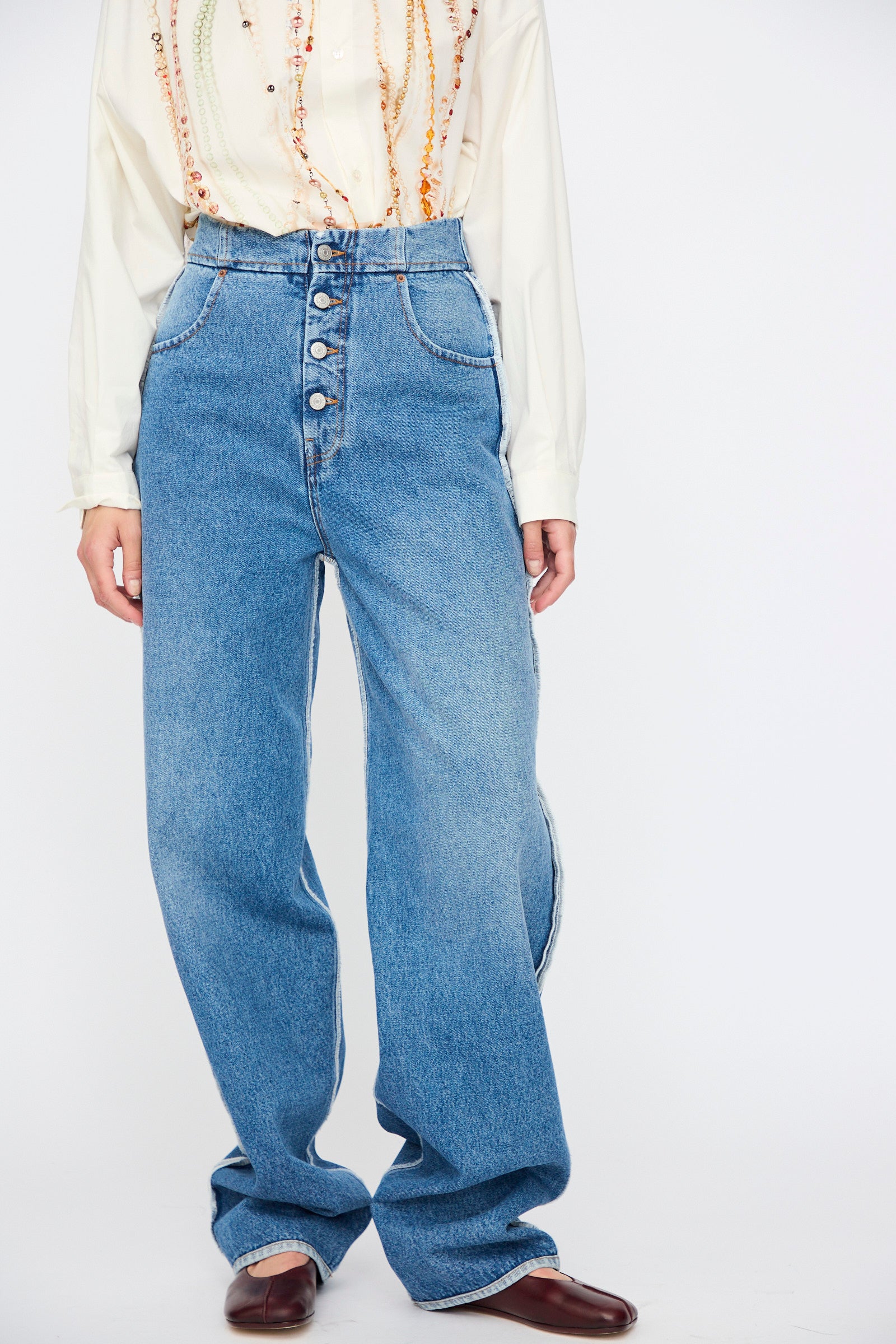 A person wears MM6 5 Pocket Pants in Medium Blue with a cream long-sleeve embroidered blouse and brown shoes, standing against a plain white background.