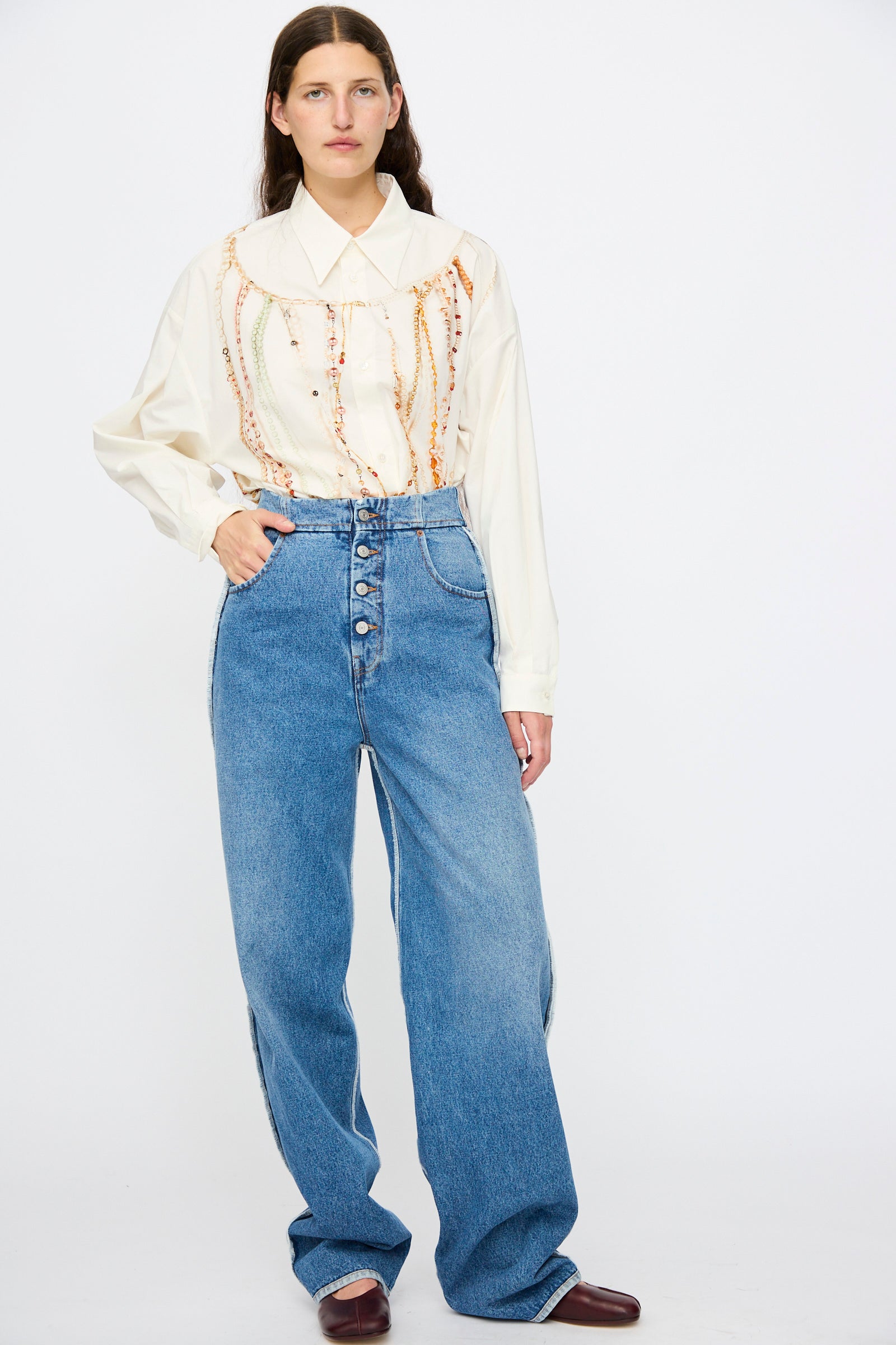 A person stands against a white background wearing a cream blouse with bead details, MM6 5 Pocket Pants in Medium Blue, and brown shoes.