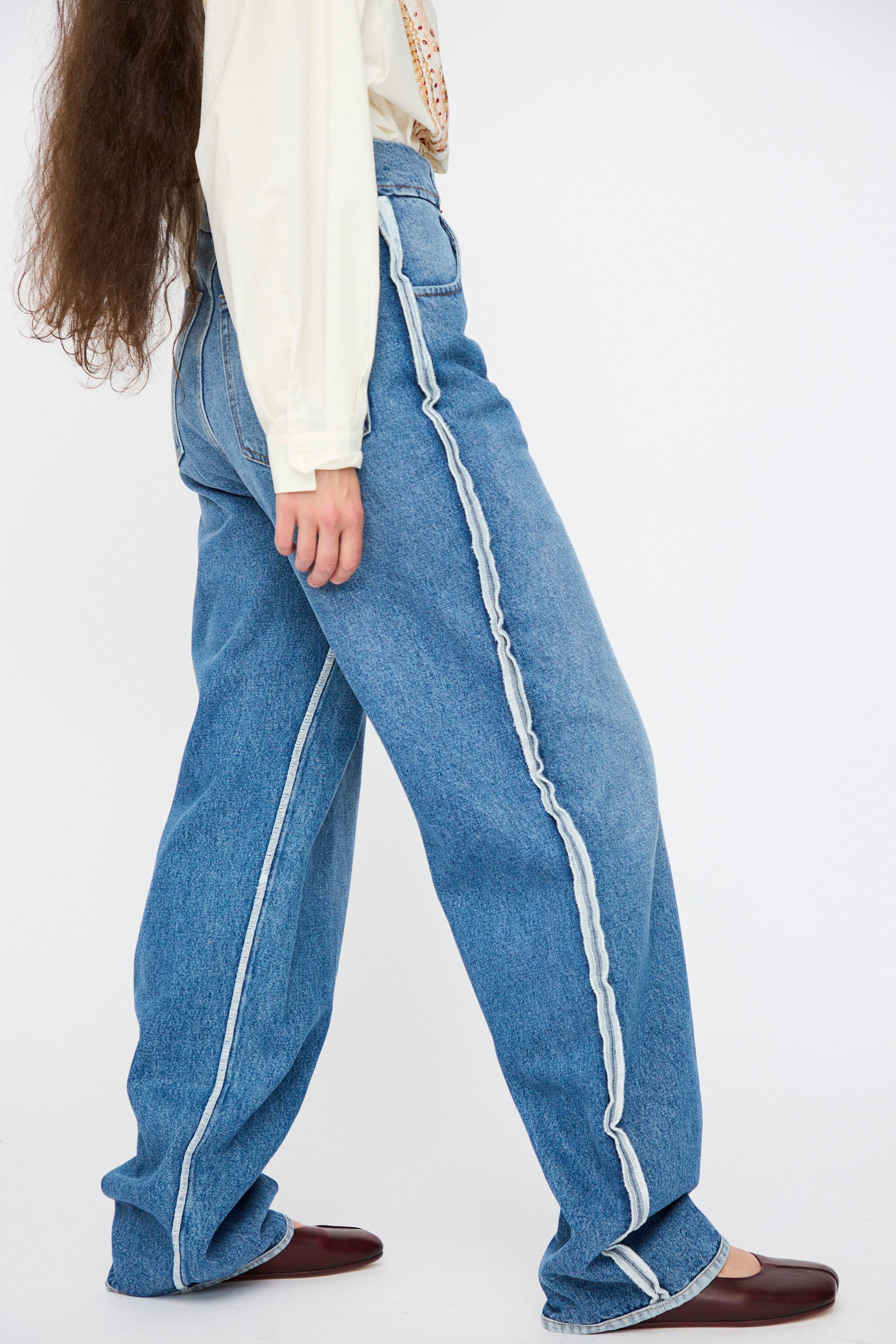 Model shown from the side in MM6 5 Pocket Pants in Medium Blue, featuring loose, wide legs with exposed white side seams, paired with a cream long-sleeve shirt and dark brown flat shoes against a plain background.