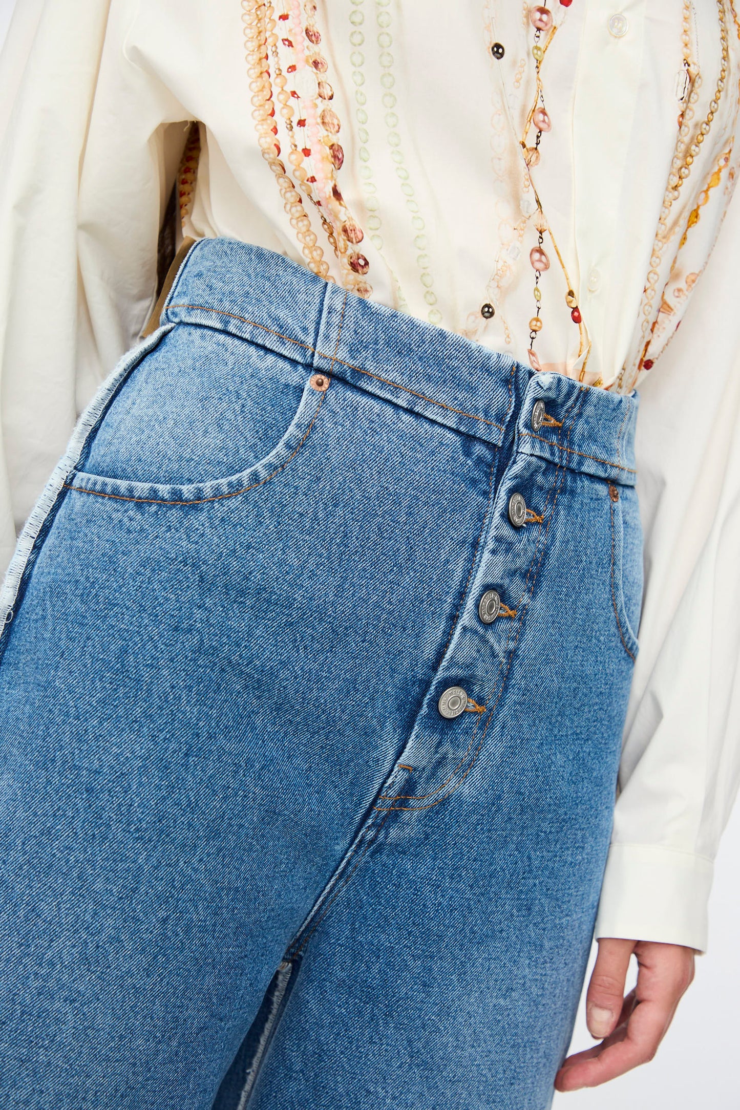 A person wearing MM6 5 Pocket Pants in Medium Blue denim styled with a cream button-up shirt and layered beaded necklaces.