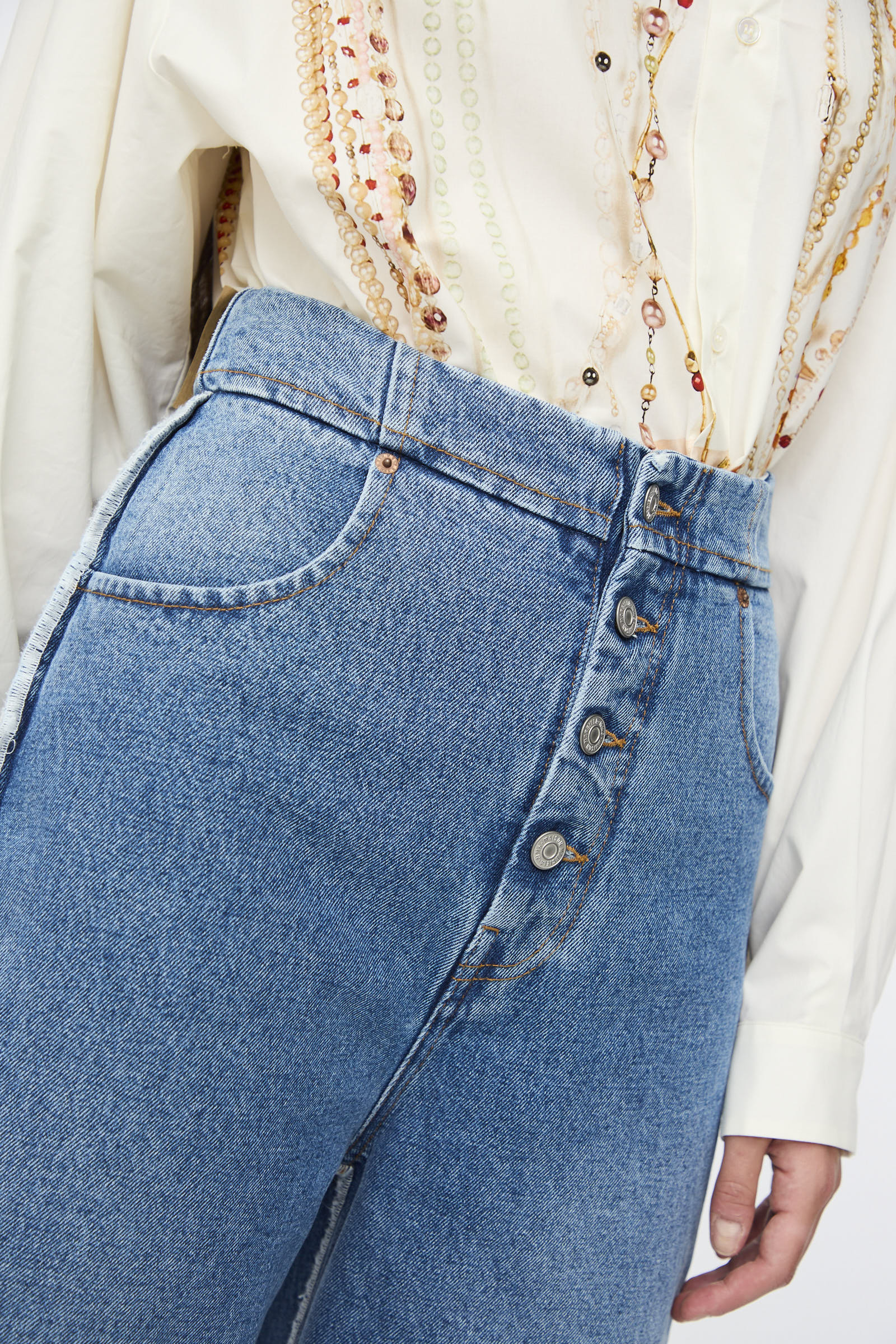 A person wearing MM6 5 Pocket Pants in Medium Blue denim styled with a cream button-up shirt and layered beaded necklaces.
