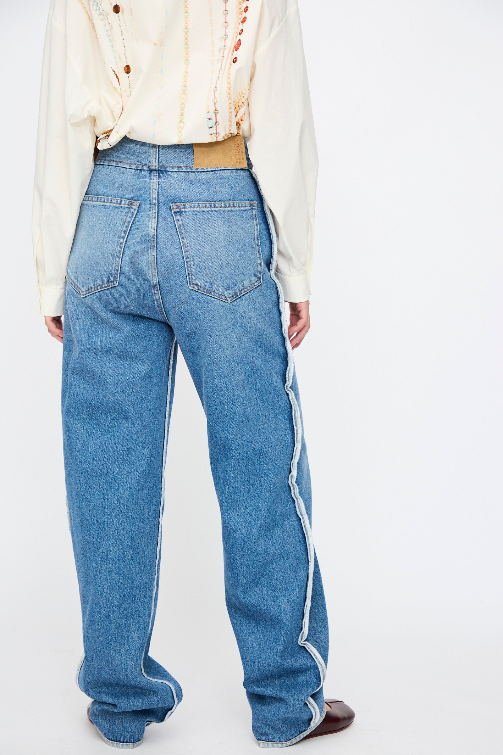 A person wearing MM6 5 Pocket Pants in Medium Blue—high-rise, wide-leg denim jeans with back pockets and white side trim—paired with a cream shirt, shown from the back.